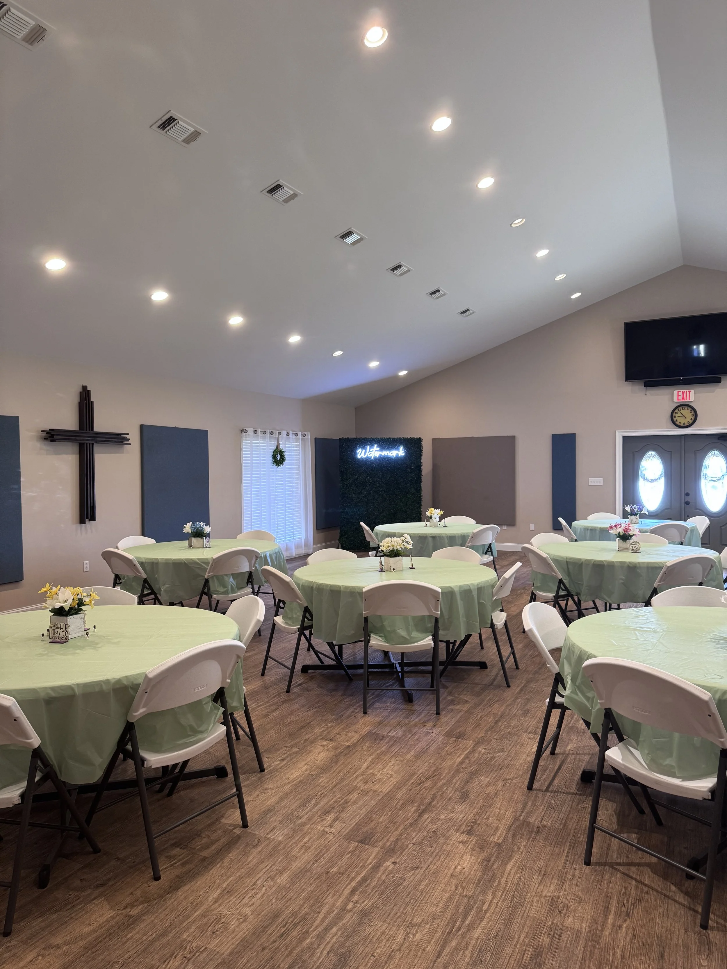 A decorated banquet hall with round tables covered in light green tablecloths and white folding chairs. Each table has small floral centerpieces, and there is a wall with decorative soundproof panels, a neon sign, and a door with oval glass panels. T