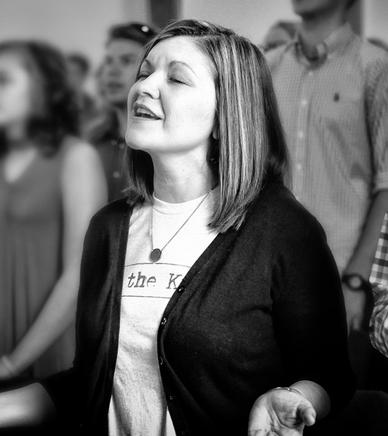 A woman with shoulder-length hair, eyes closed, and a peaceful expression, standing among a group of people.