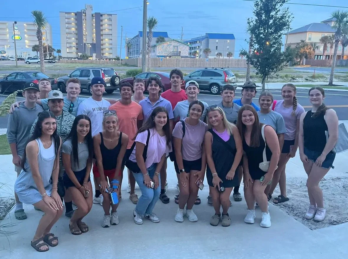A group of young people, both boys and girls, posing outdoors on a sidewalk in front of parked cars with buildings and palm trees in the background.