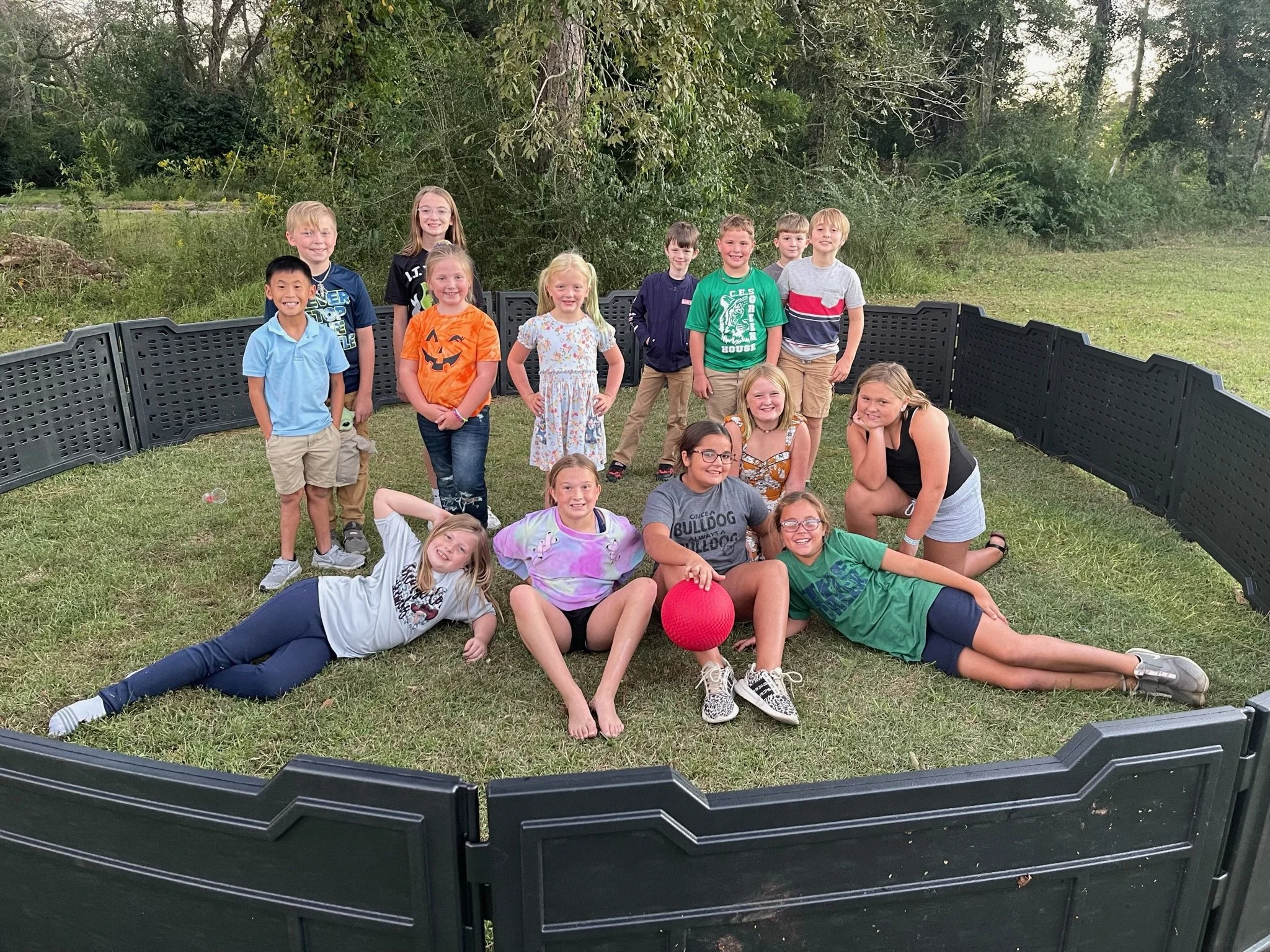 Group of children posing outdoors near a black fence with trees and bushes in the background.