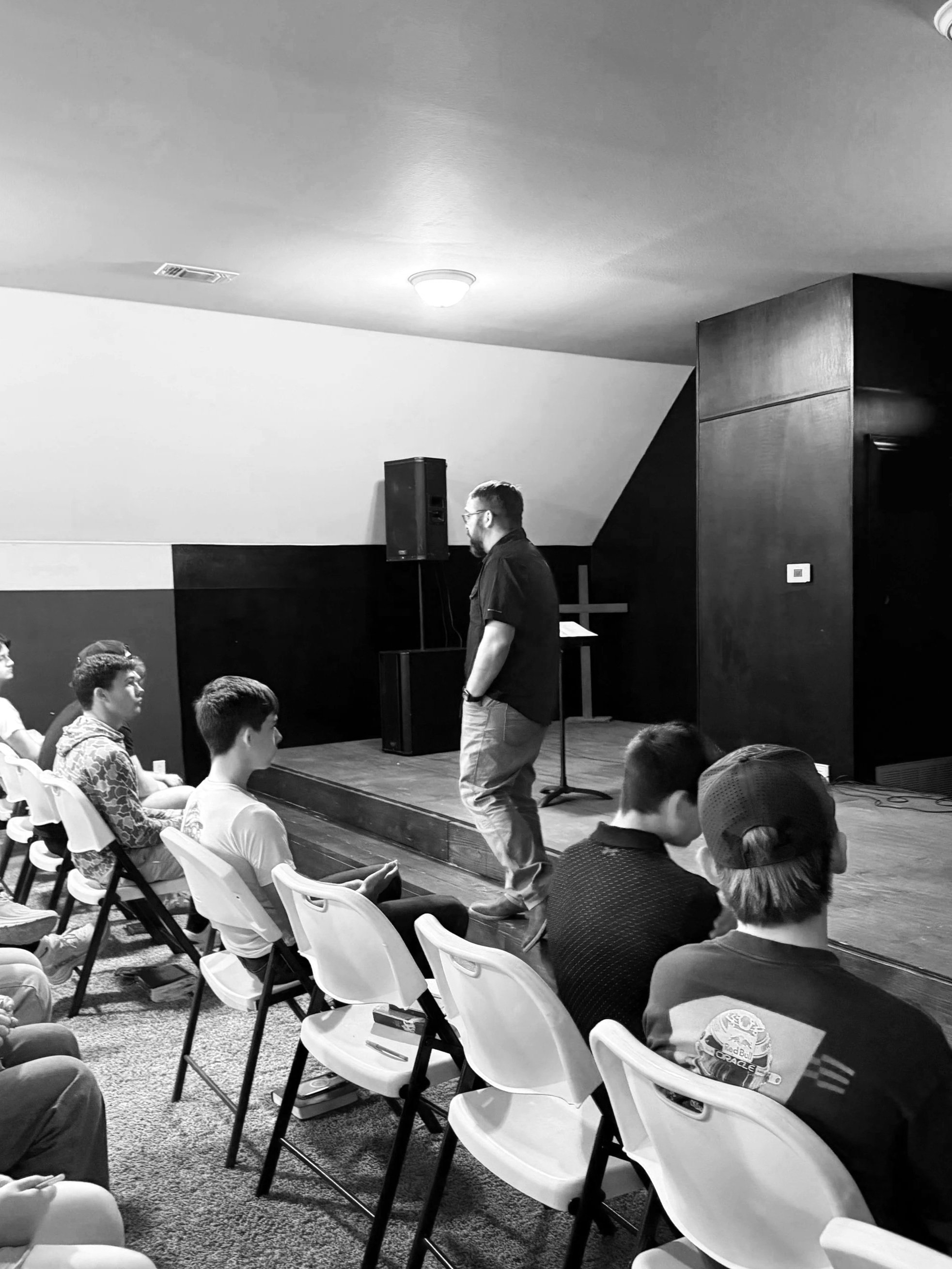 A man on stage speaking to an audience of young people in a room with a high ceiling and wood-paneled walls