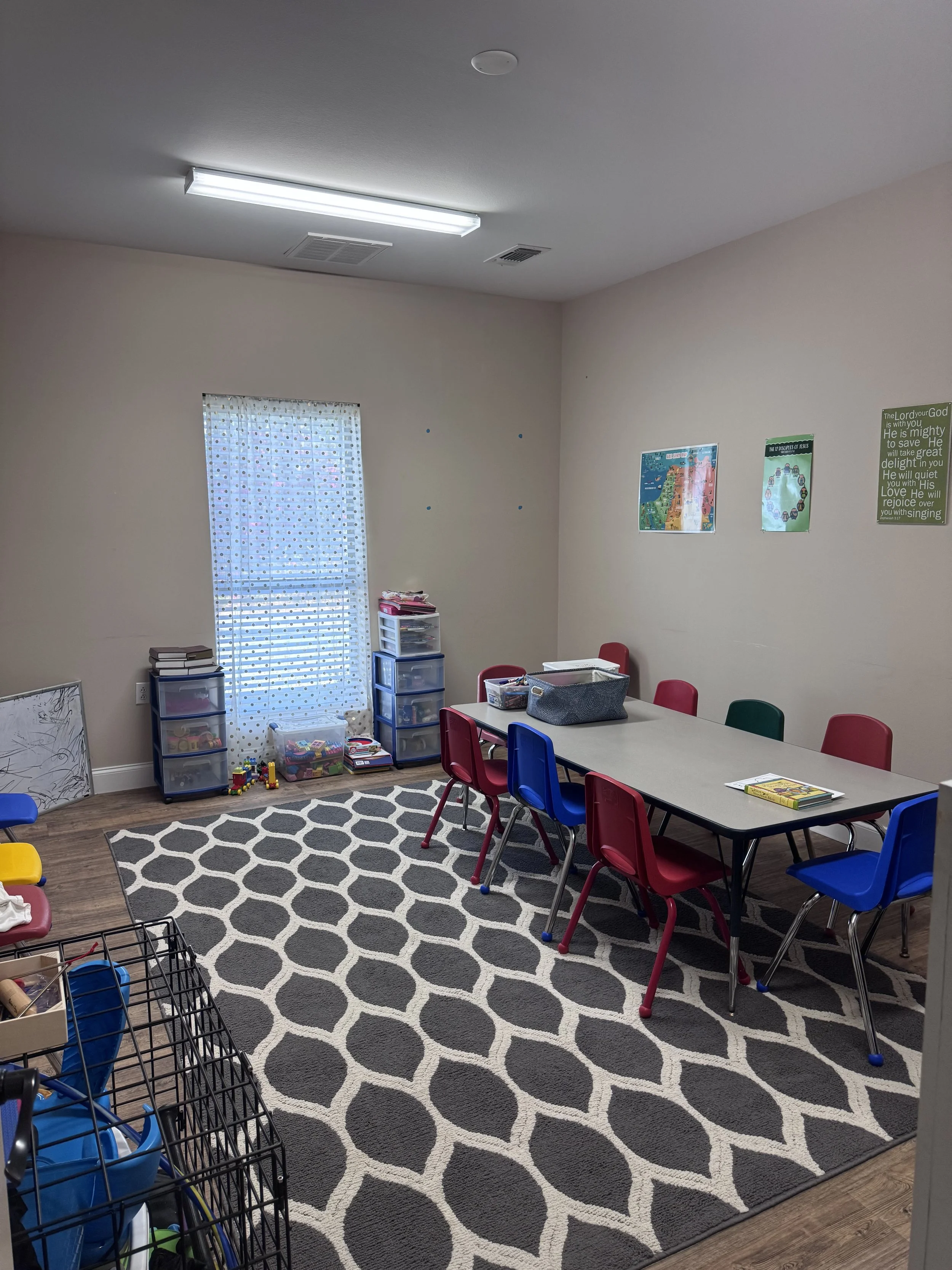 Classroom with a table and colorful chairs, a black and white patterned rug, shelves with storage bins and toys, and educational posters on the wall.