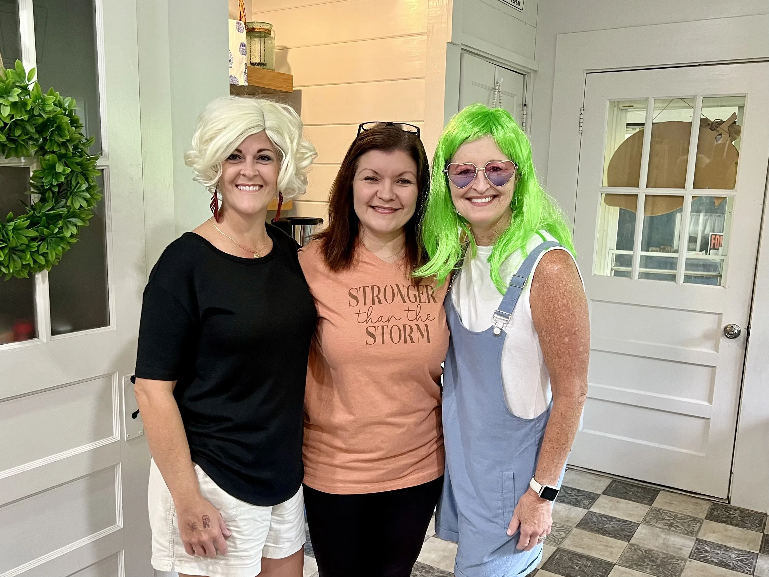 Three women standing side by side inside a house, smiling at the camera. The woman on the left has short, curly platinum blonde hair, and is wearing a black top. The woman in the middle has shoulder-length brown hair, glasses on her head, and is wear