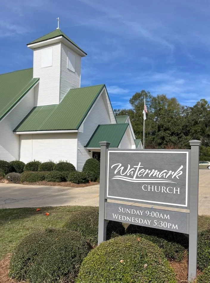 White church with green roof, shrubs in front, and a sign that reads Watermark Church with service times Sunday 9:00 AM and Wednesday 5:30 PM, under a partly cloudy blue sky.