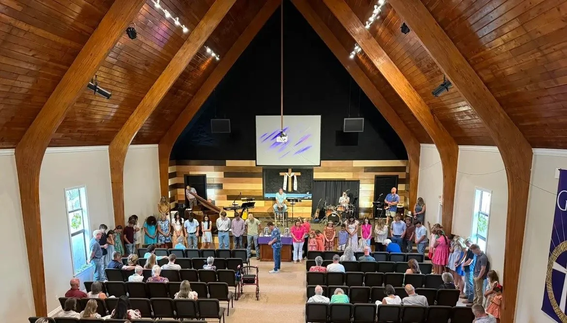 People gather inside a church sanctuary for a worship service or event, with a band on stage and a cross on the wall behind them.