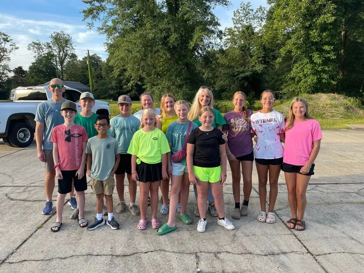 Group of children and two adults posing outside on a paved area with trees in the background.