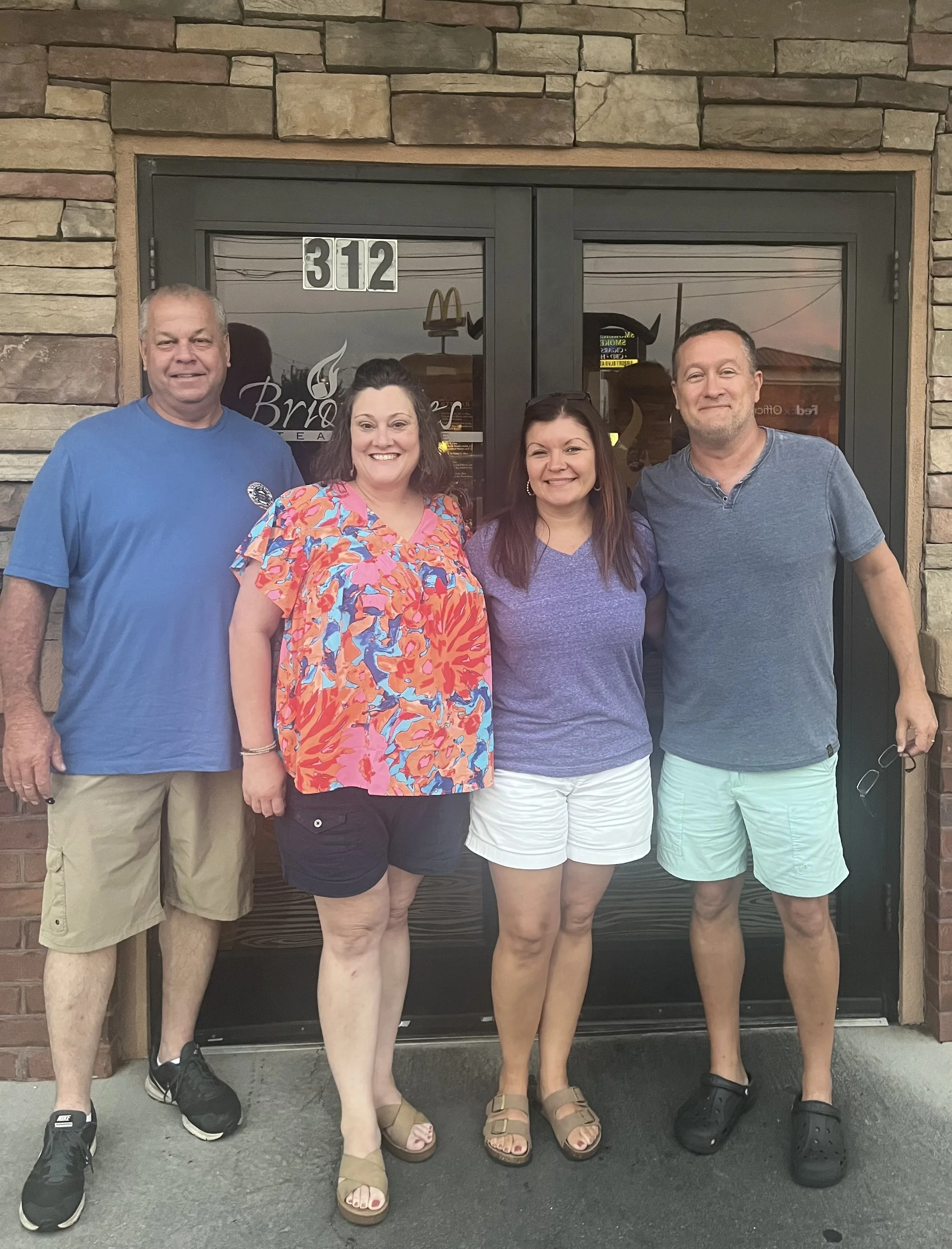 Four people standing in front of a building with the address 312 and a glass door with a logo that says Bri. They are smiling and dressed casually. The building has a brick exterior.