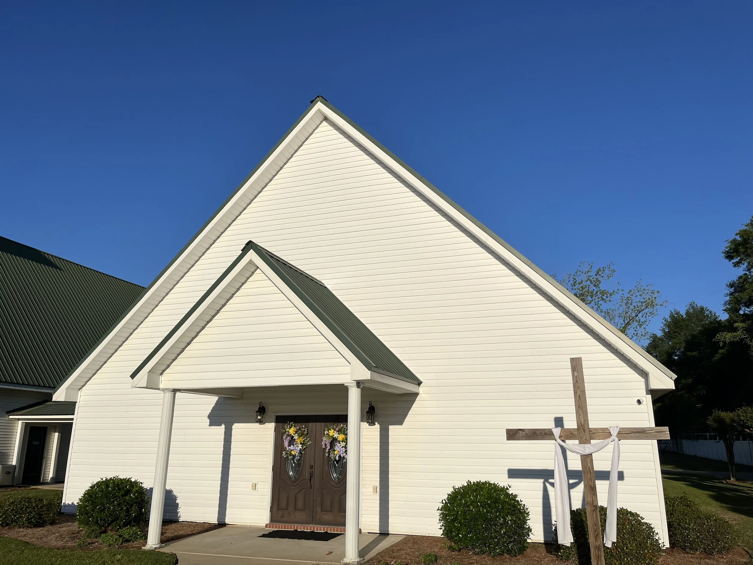 White church with pointed roof, decorated for a wedding, with a wooden cross with white cloth, and floral arrangements, against a blue sky with trees.