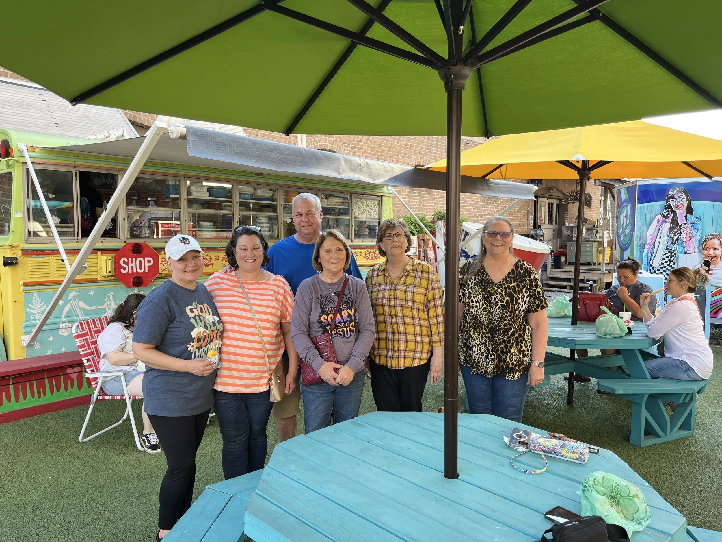 Group of six women standing together outdoors under large green and yellow umbrellas, with a colorful food truck and other people in the background, smiling and posing for the photo.