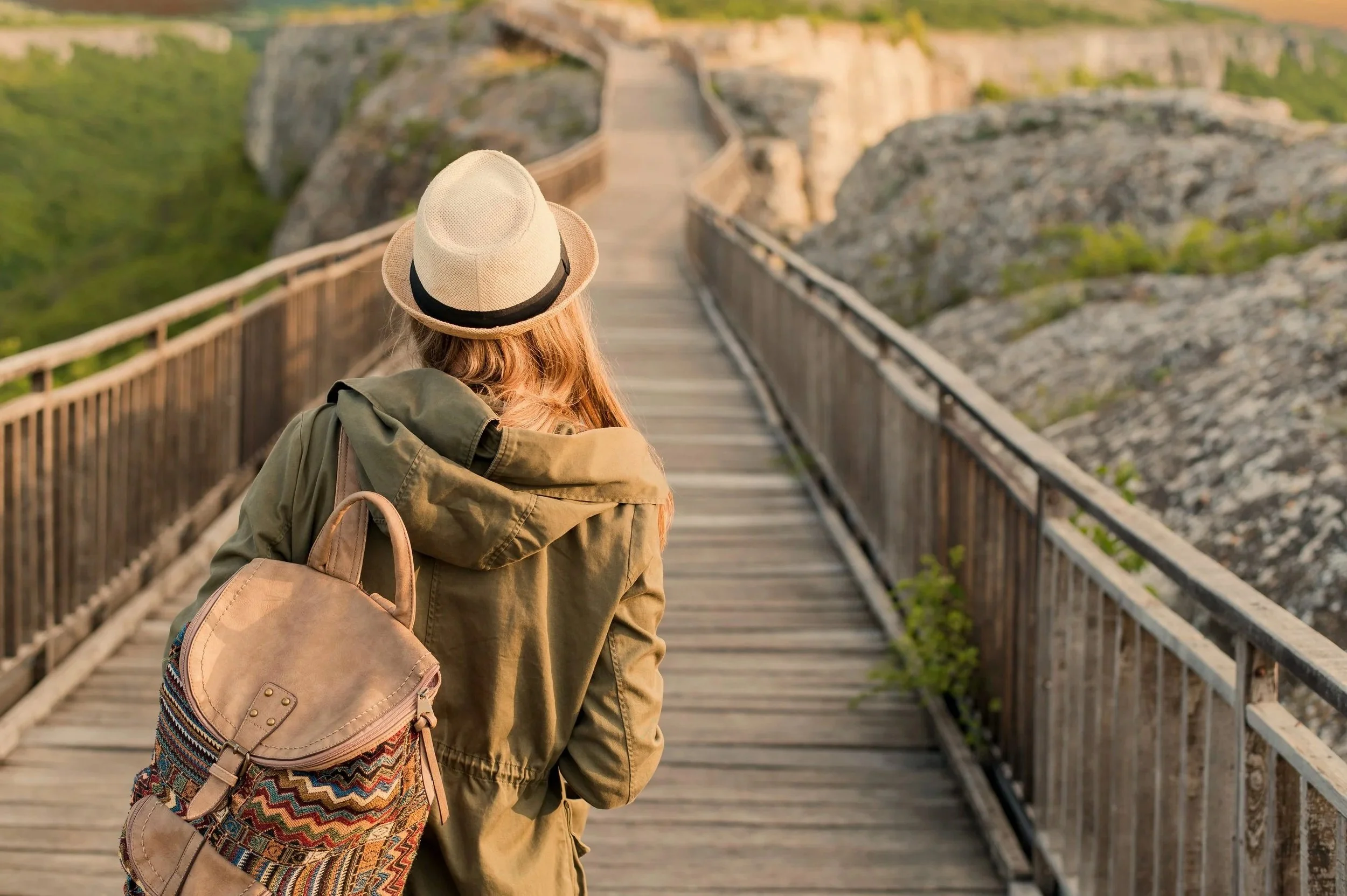 Woman with long hair, wearing a hat and carrying a backpack, walking on a wooden bridge over a rocky landscape with greenery in the background.