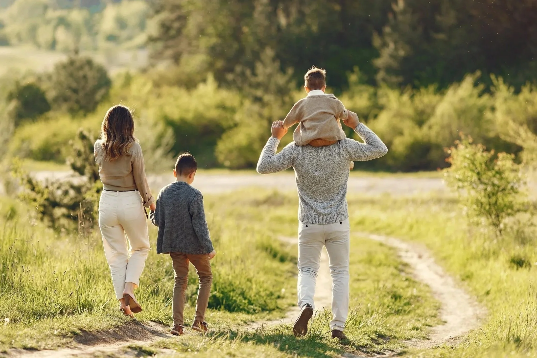 A family walking outdoors along a grassy path in a green, wooded area during daytime. A man carries a young boy on his shoulders, while a woman and a young boy walk beside him.