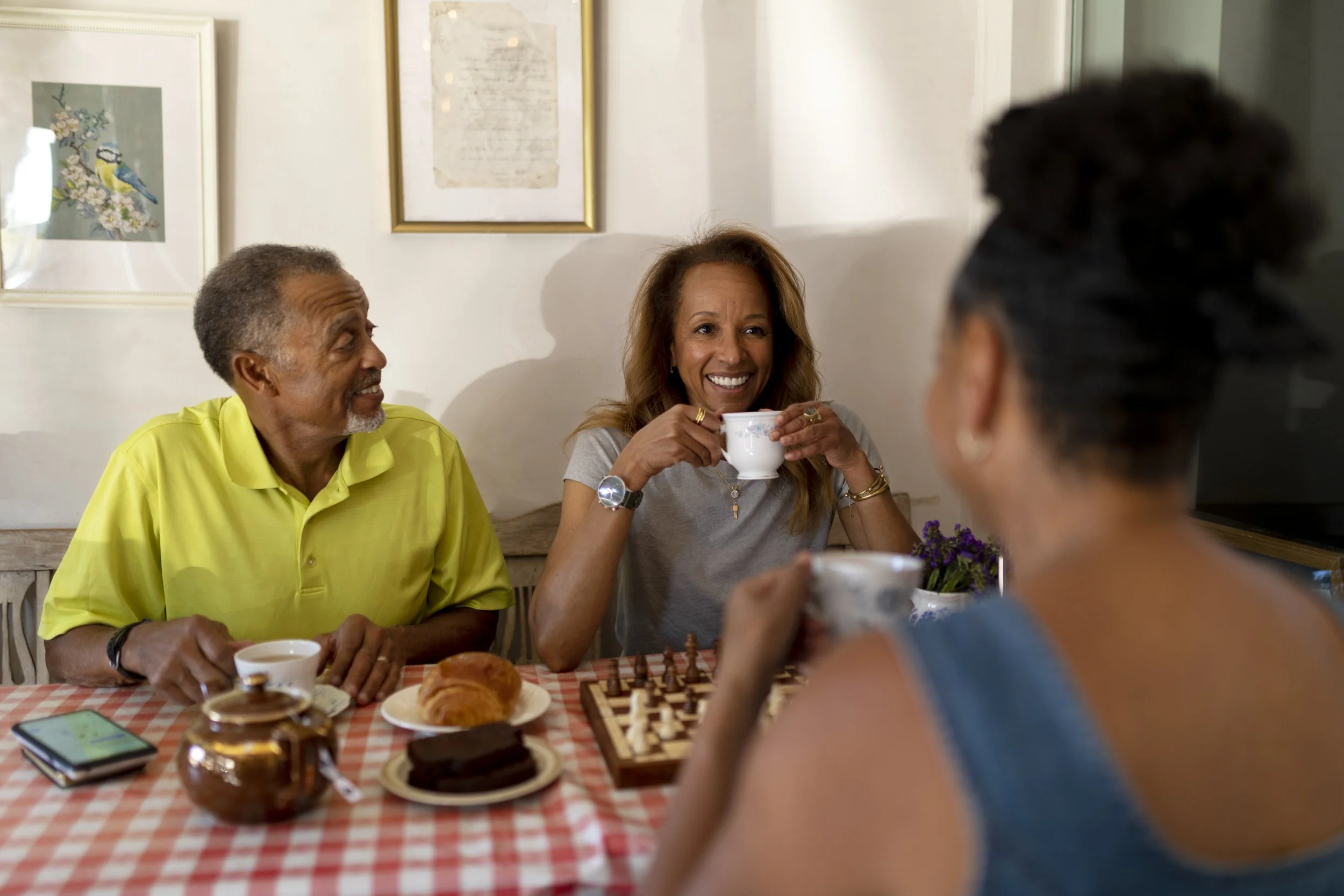 A group of four people sitting around a table having tea and playing chess. The table is covered with a red and white checkered tablecloth, with baked goods and a chessboard on it. The background shows framed artwork and a white wall. The bearded man in a yellow shirt and the woman with long hair holding a teacup are visible, while two other women are partially visible.