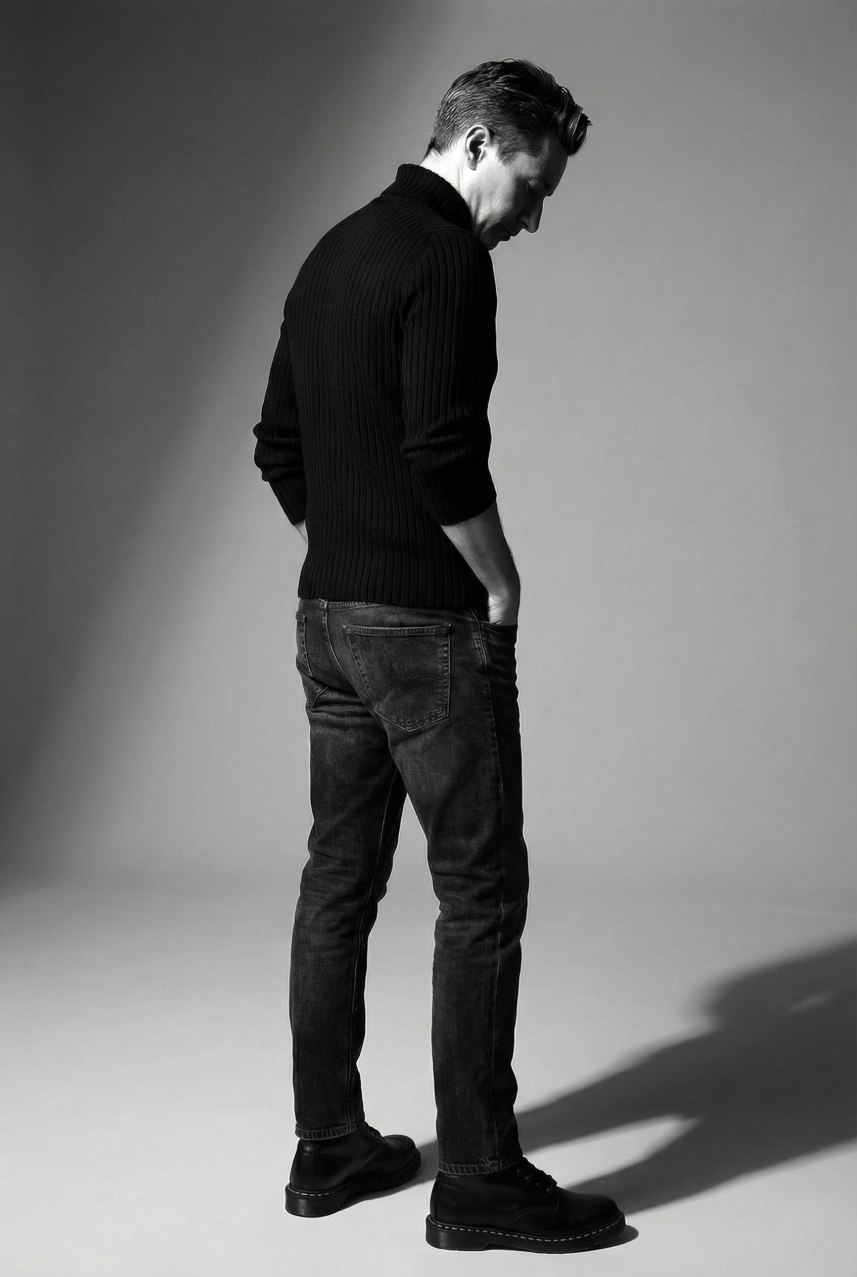 Black and white photograph of Merlin Zuni in dark jeans, black boots, and a black ribbed sweater, standing with his head slightly bowed and hands behind his back on a plain background with shadows.