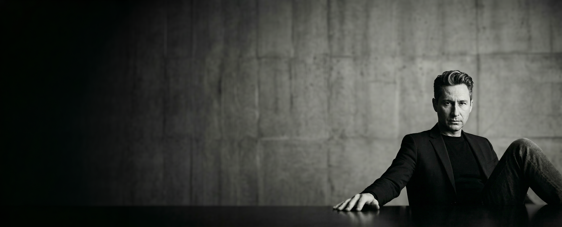 Merlin Zuni sitting against a concrete wall in a black blazer and black shirt looking stoic, arm resting on the table in a black and white portrait.