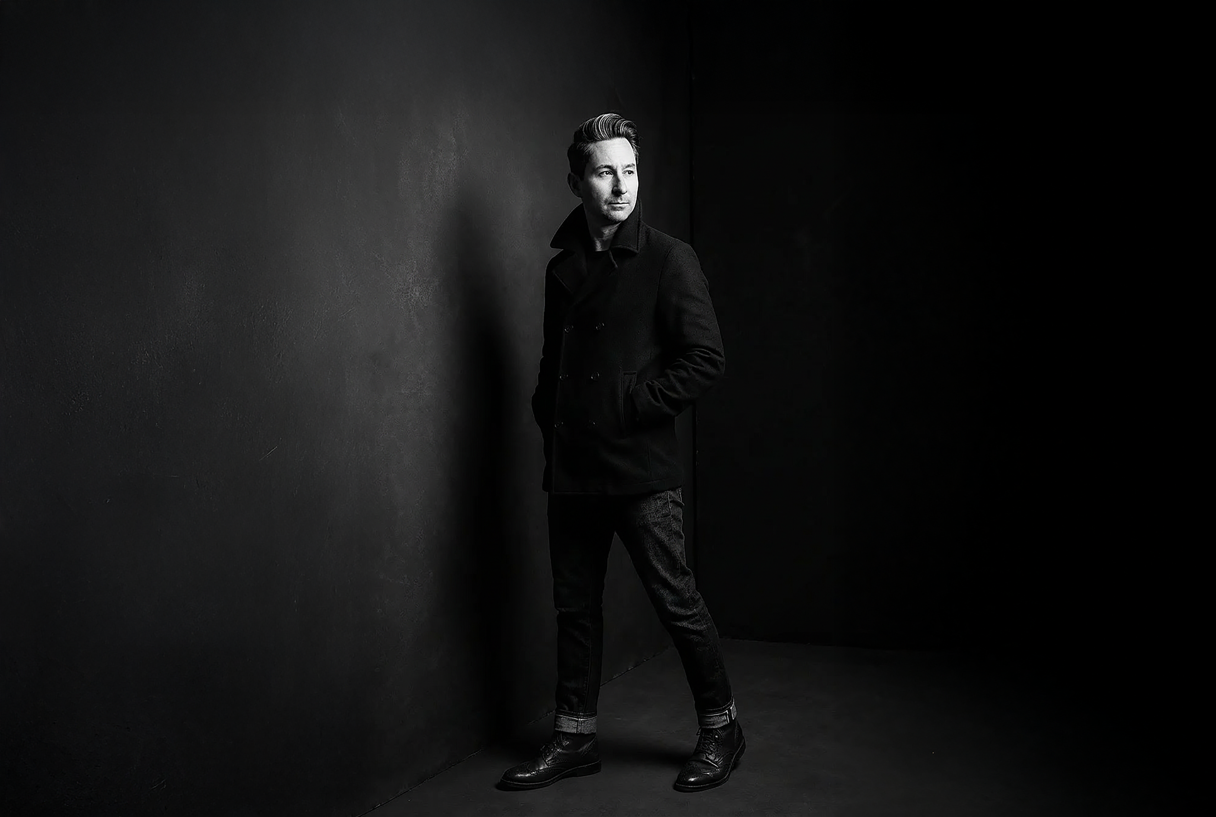 A portrait photograph of Merlin Zuni standing against a dark wall, looking to the side, wearing a black peacoat and dark pants with boots, in a black and white photograph.