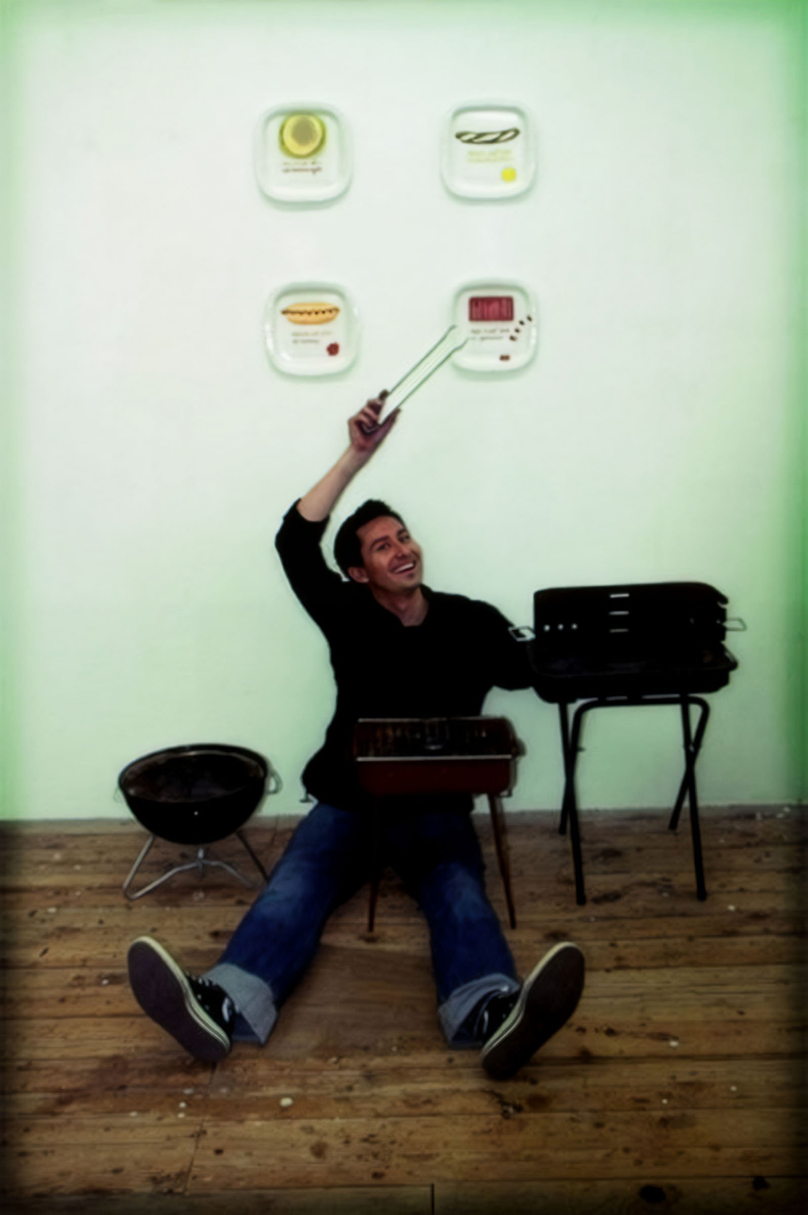 Art Title - Grill Teller, Merlin Zuni, photo of Merlin Zuni sitting next to gallery installation of trompe l'oeil bbq plates with painted food.