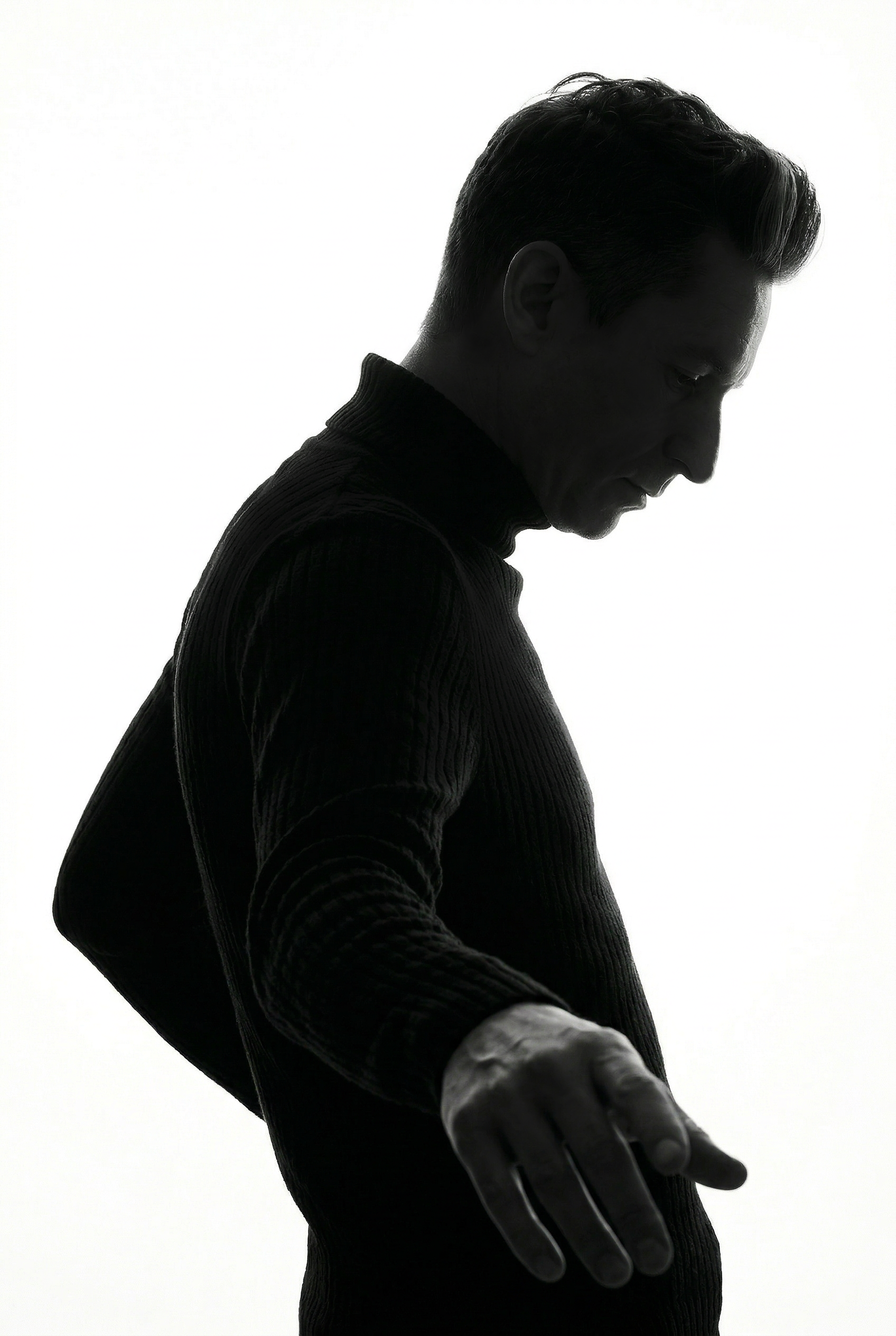 Silhouette of Merlin Zuni in a black turtleneck sweater standing against a white background, with focus on his side profile and outstretched hand.