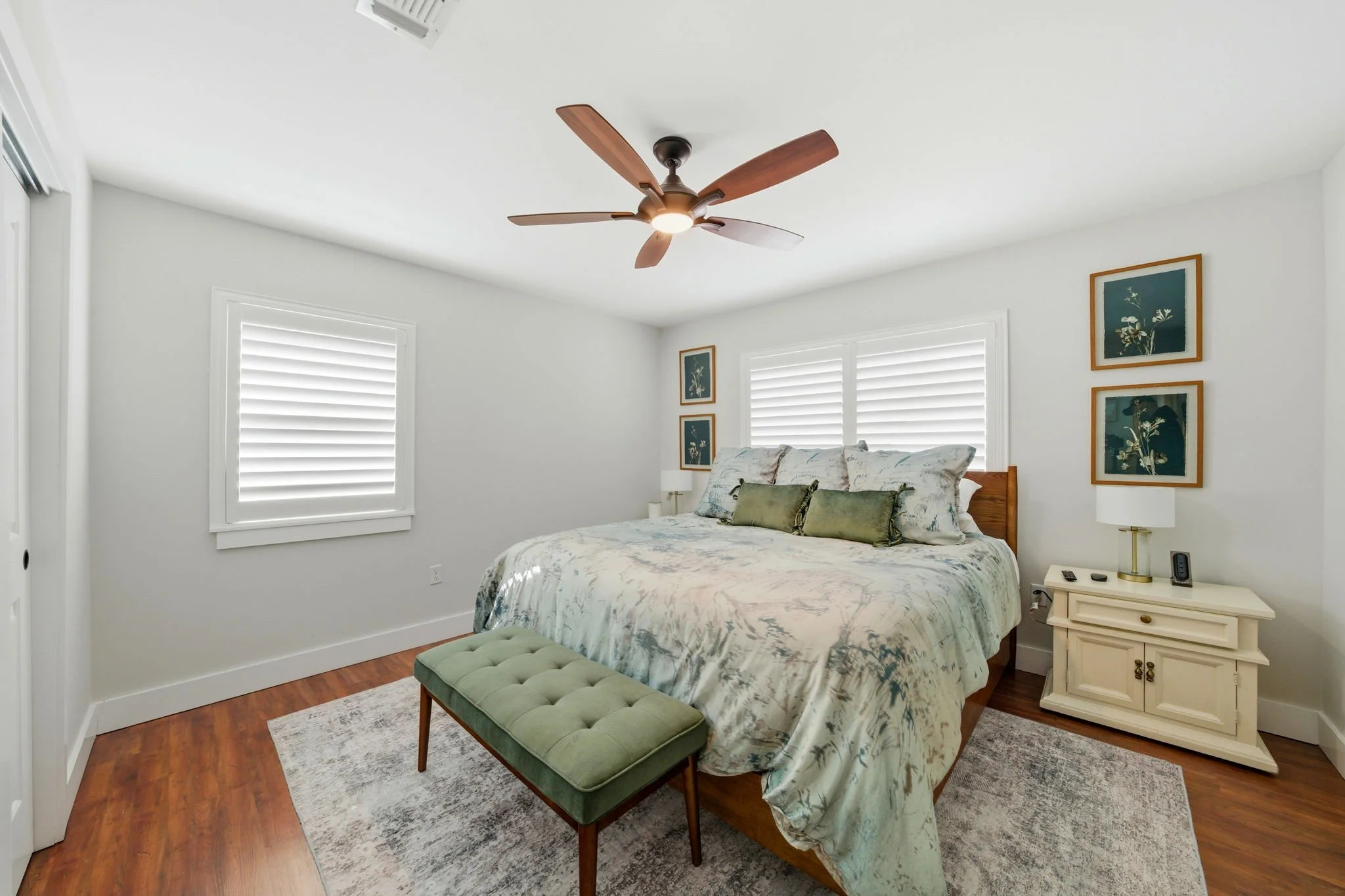 A bedroom with white walls, a large bed with a floral duvet, green throw pillows, and a wooden headboard. There is a green bench at the foot of the bed, a white nightstand with a lamp and electronics, and framed botanical artwork on the wall. Wooden flooring and white blinds on the windows.