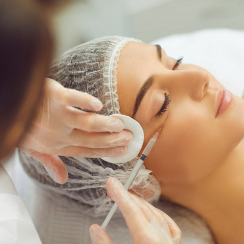 A woman receiving a beauty treatment on her eye area, with a practitioner using a small brush and a cotton pad.