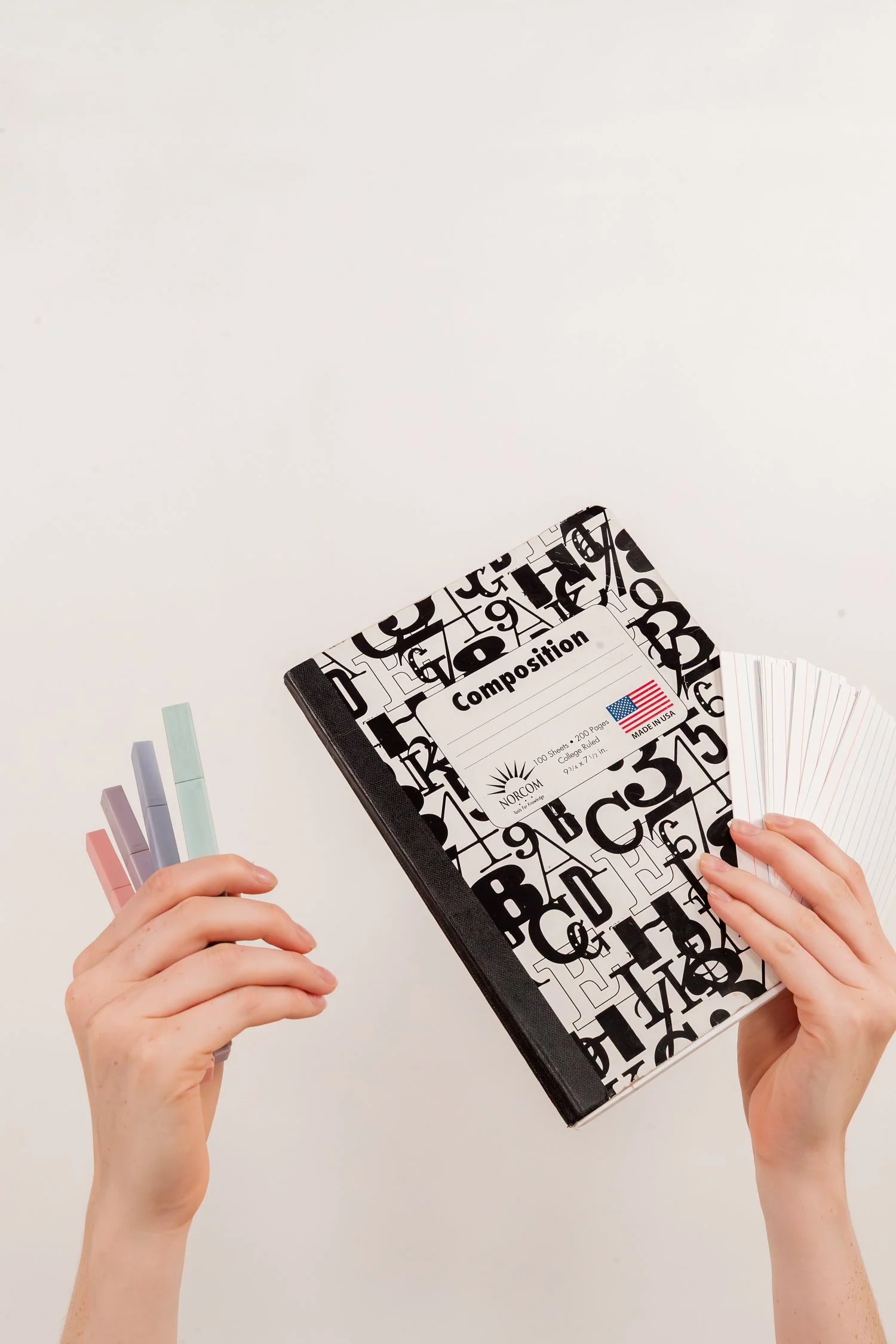 Hands holding a composition notebook, blank index cards, and pastel highlighters on a white background