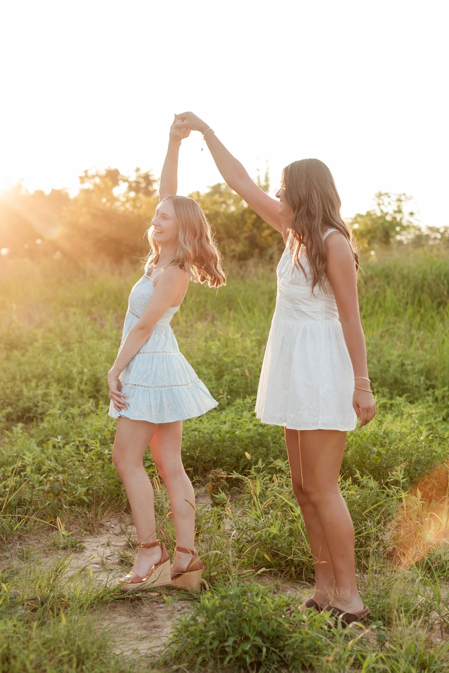 Senior in a light blue sundress spinning under a friend's raised hand in a grassy field, bestie senior pictures in Bolivar, MO