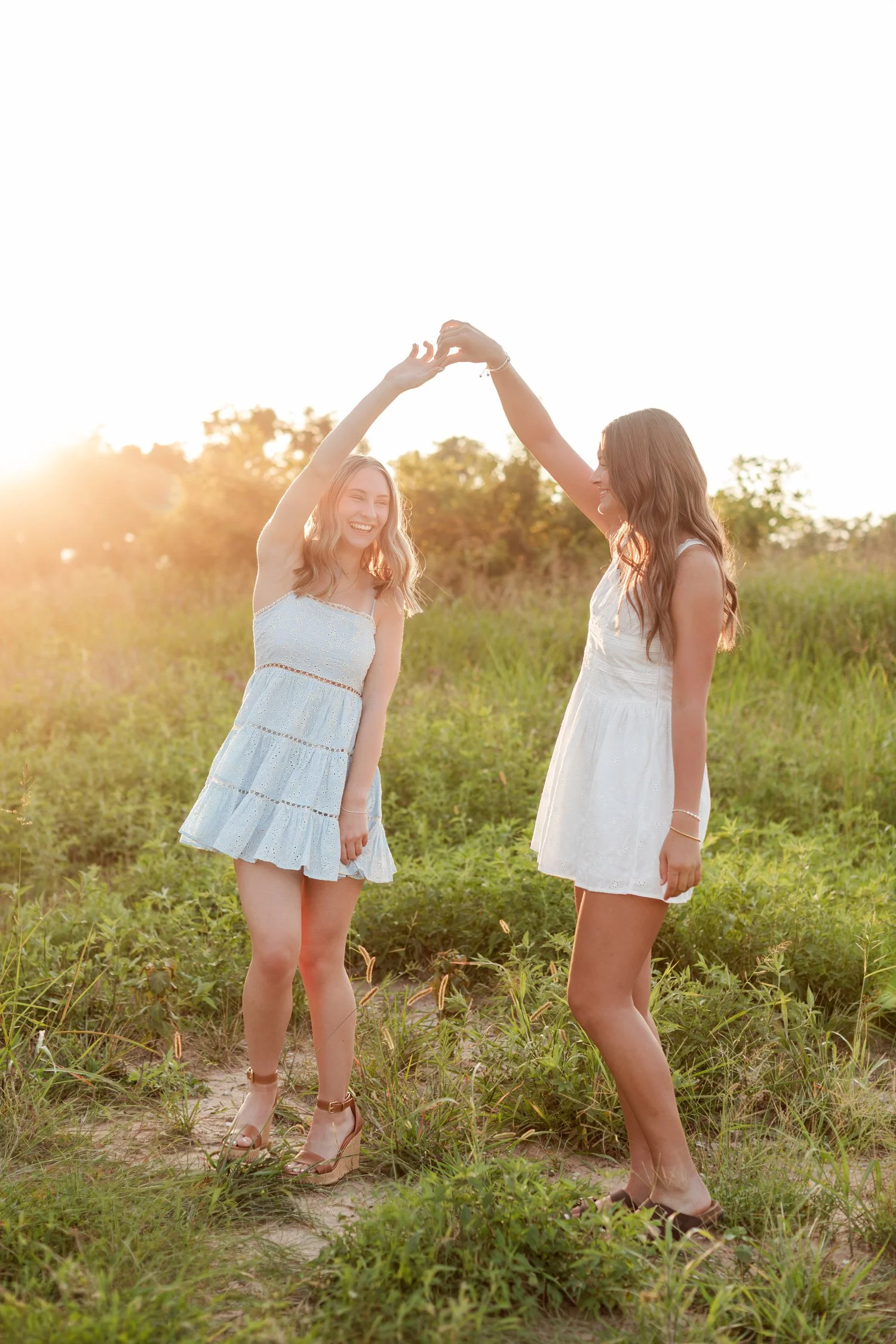 Two seniors holding raised hands in an arch and laughing in a sunlit field, bestie senior pictures in Bolivar, MO
