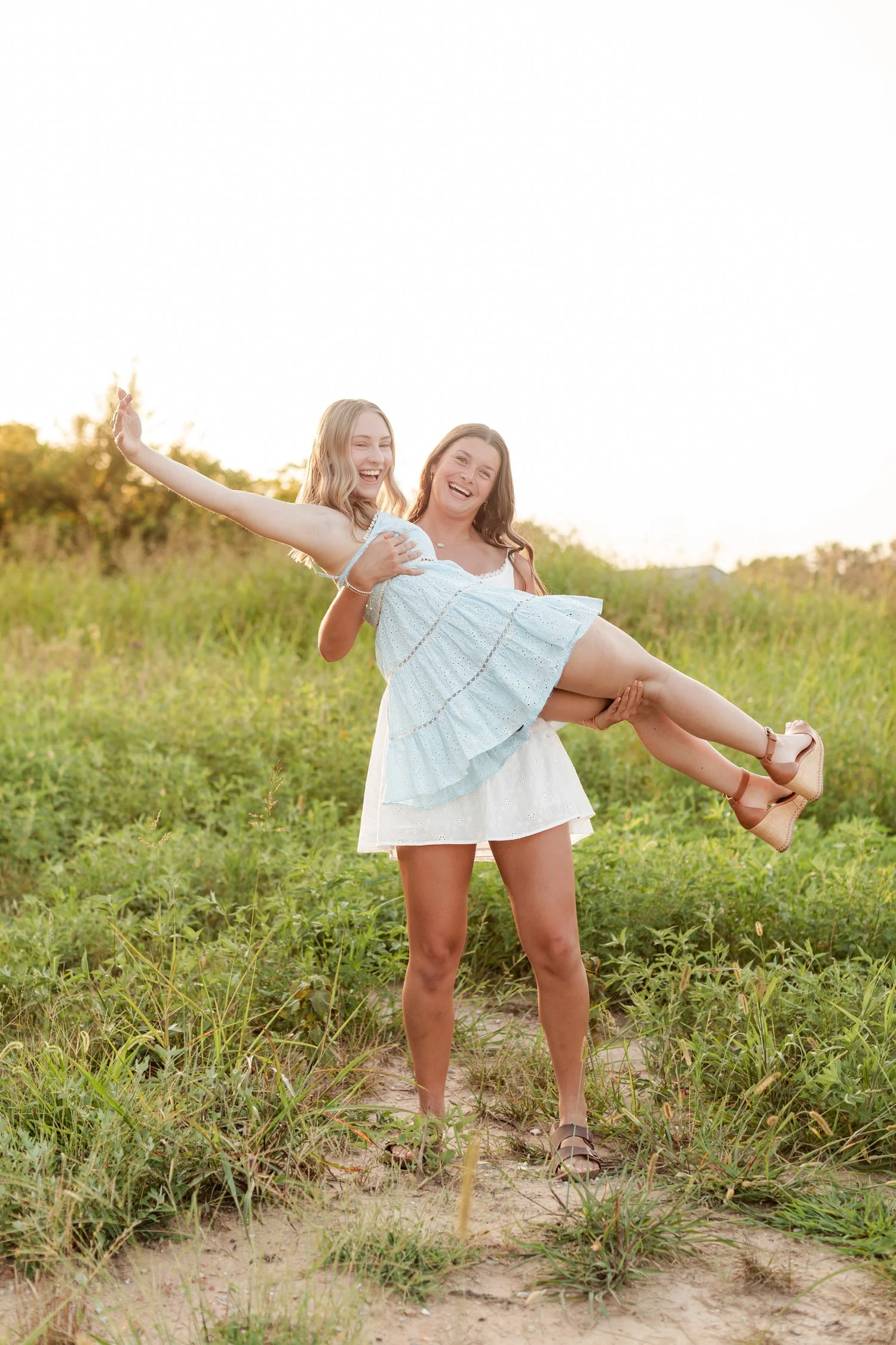 Senior in a white sundress lifting a friend in a light blue dress off the ground, bestie senior pictures in Bolivar, MO