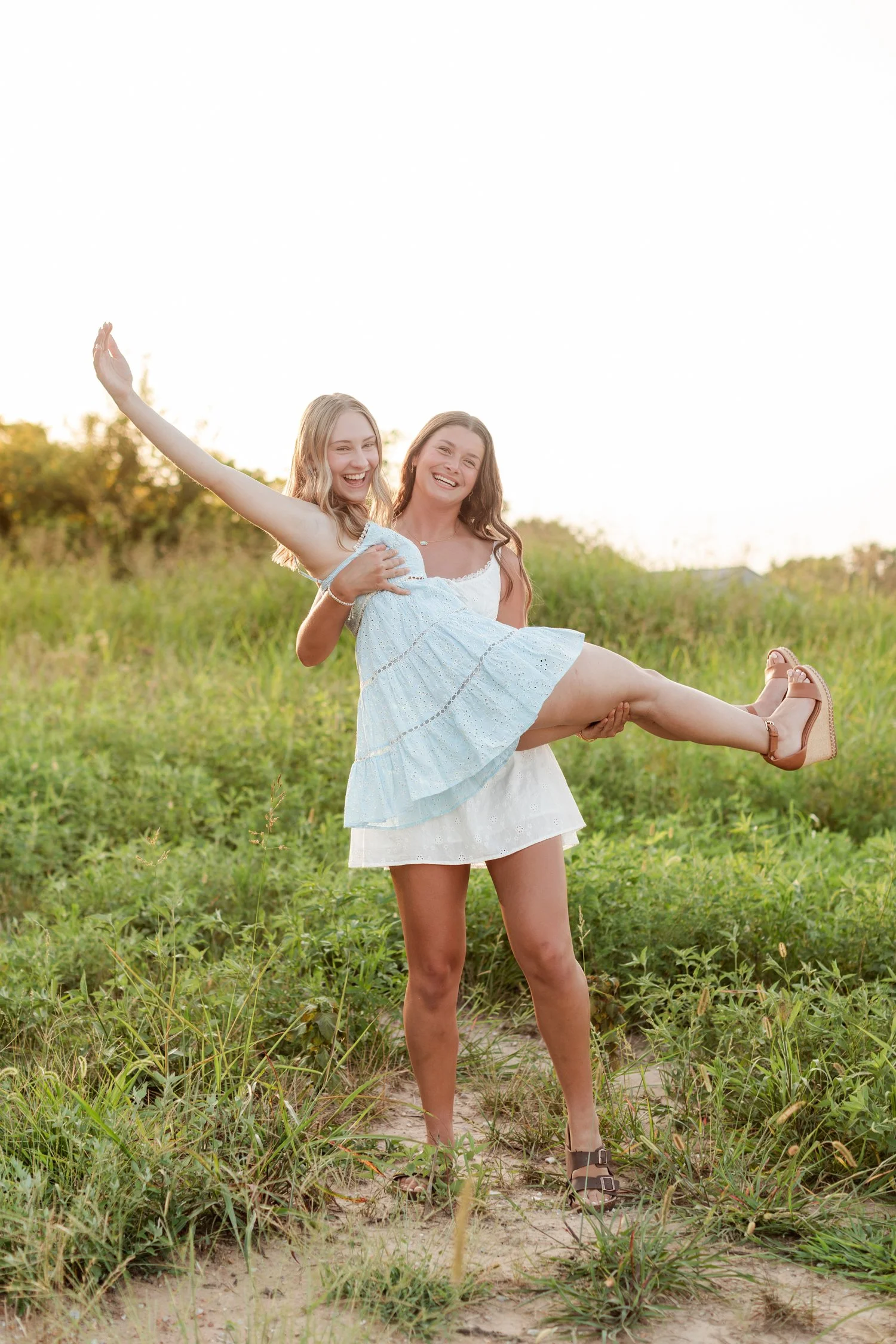 Senior in a light blue dress kicking one leg out while held by a friend in a white dress, bestie senior pictures in Bolivar, MO