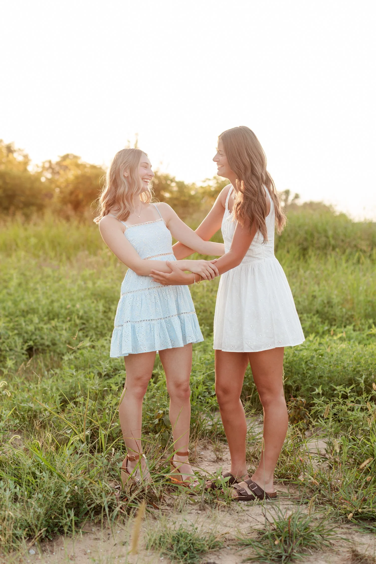 Two seniors in sundresses holding hands at arm's length in a grassy field, bestie senior pictures in Bolivar, MO