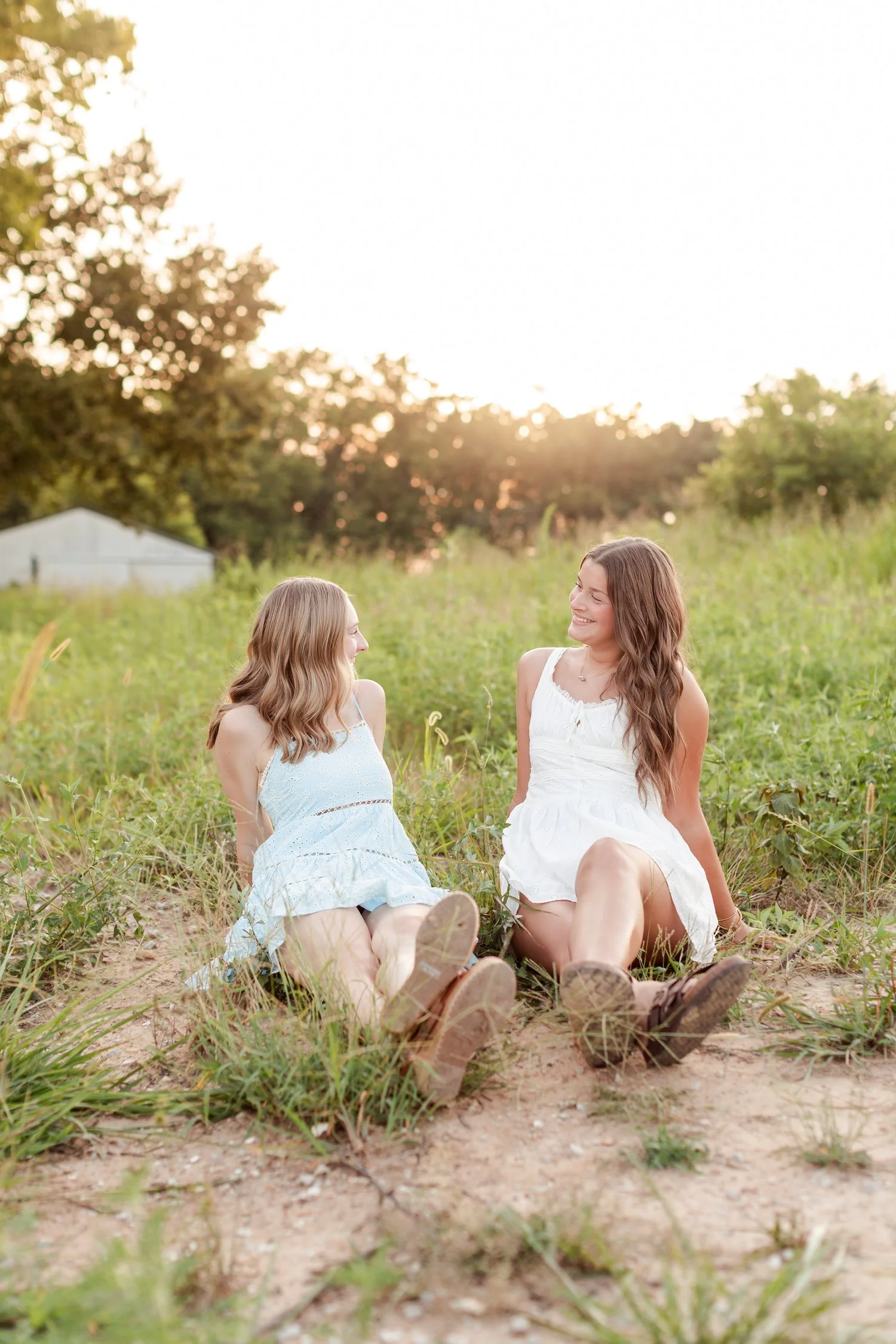 Two seniors in cowboy boots sitting on the ground a few feet apart, looking at each other, bestie senior pictures in Bolivar, MO