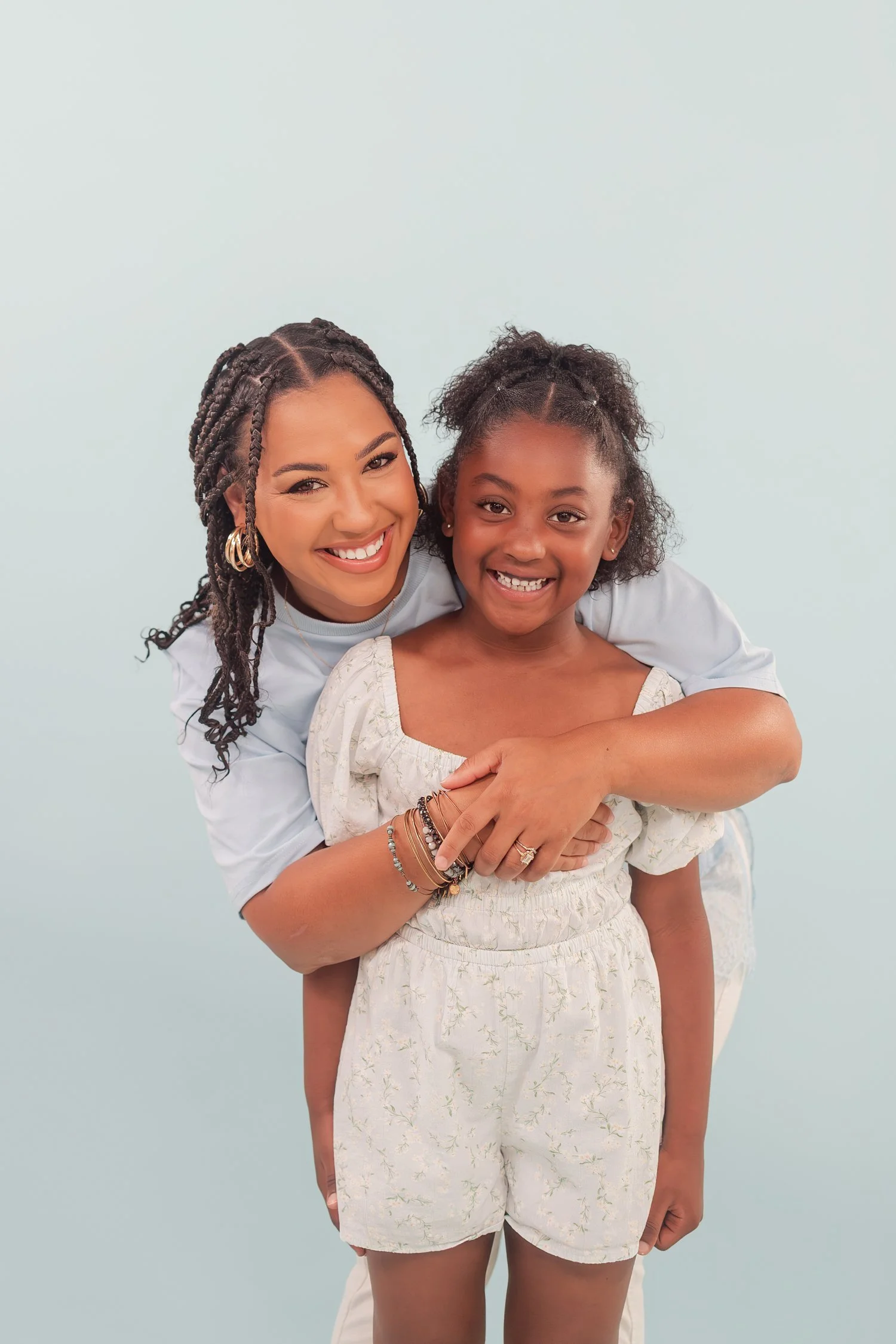 Mother in a light blue shirt hugging a smiling daughter from behind against a pale blue backdrop, mother daughter photos in Bolivar, MO