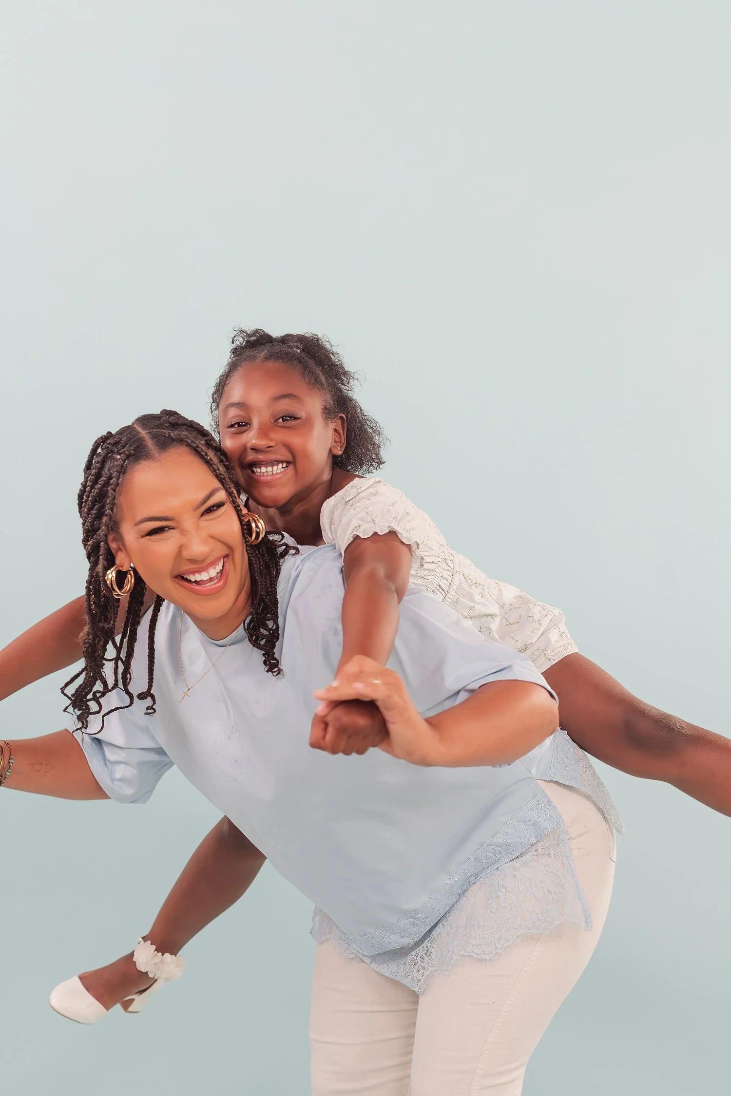 Daughter riding piggyback with arms out while mother laughs against a pale blue backdrop, mother daughter photos in Bolivar, MO
