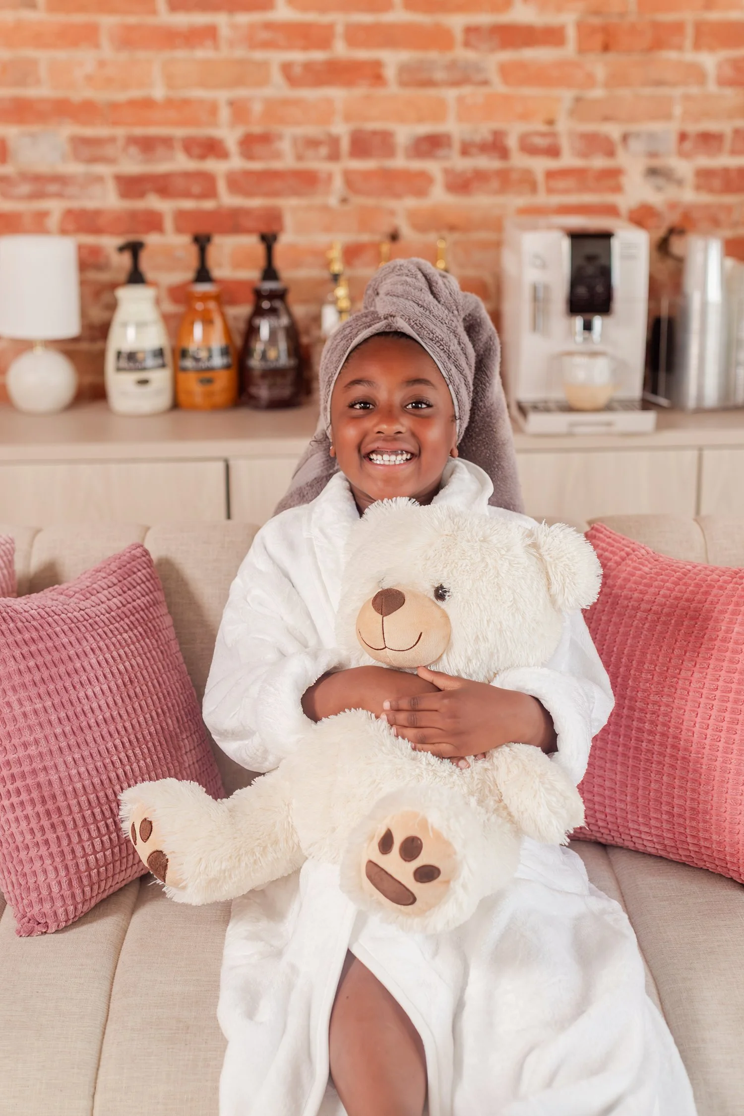 Daughter in a white robe and towel wrap holding a teddy bear on a couch with pink pillows, mother daughter photos in Bolivar, MO