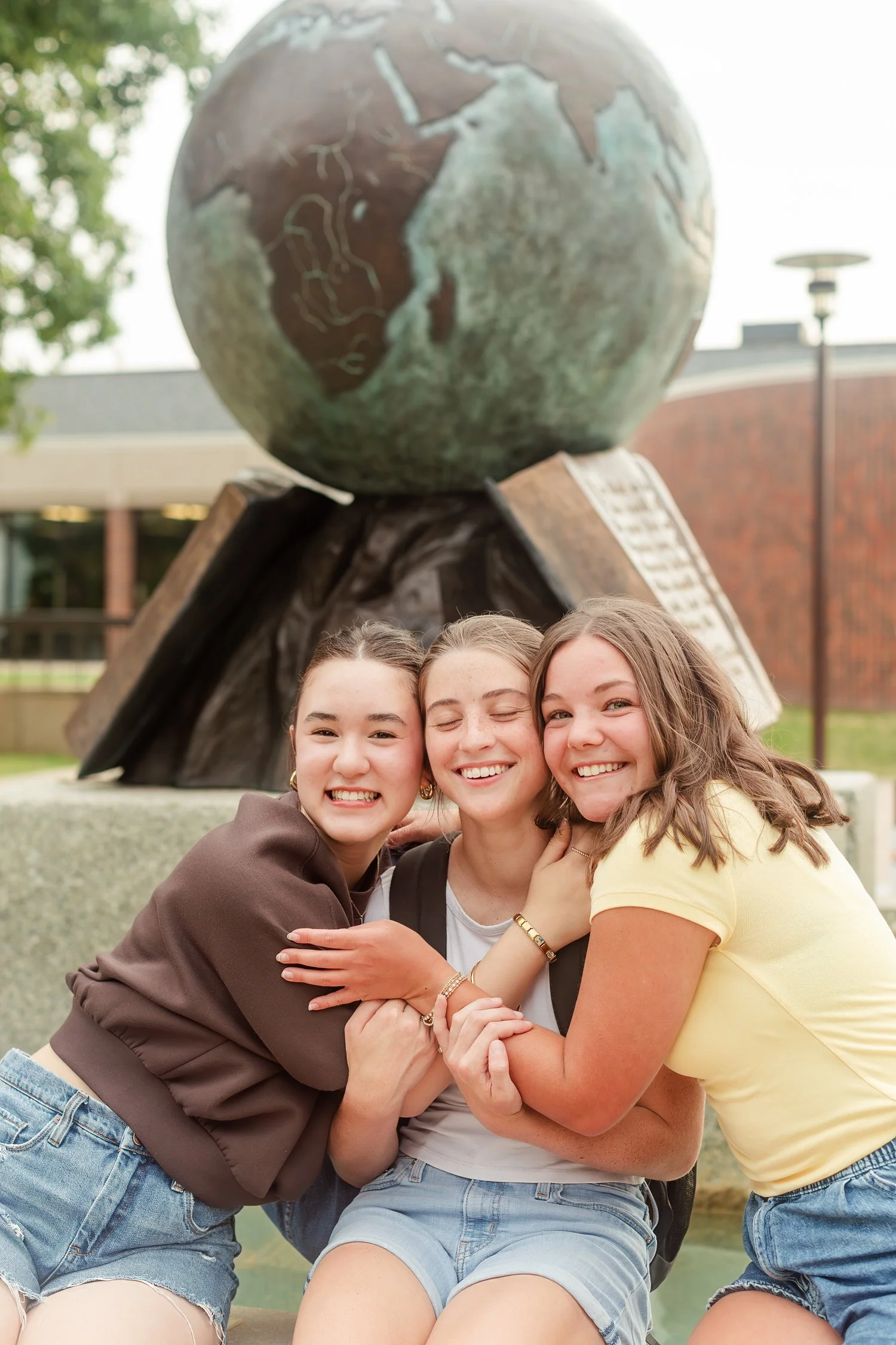 Three friends leaning together and smiling beneath a globe sculpture.