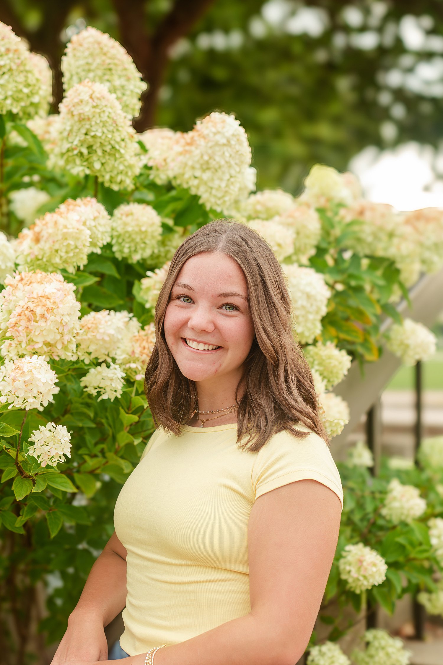 Young woman in a yellow top smiling beside white flowering shrubs.