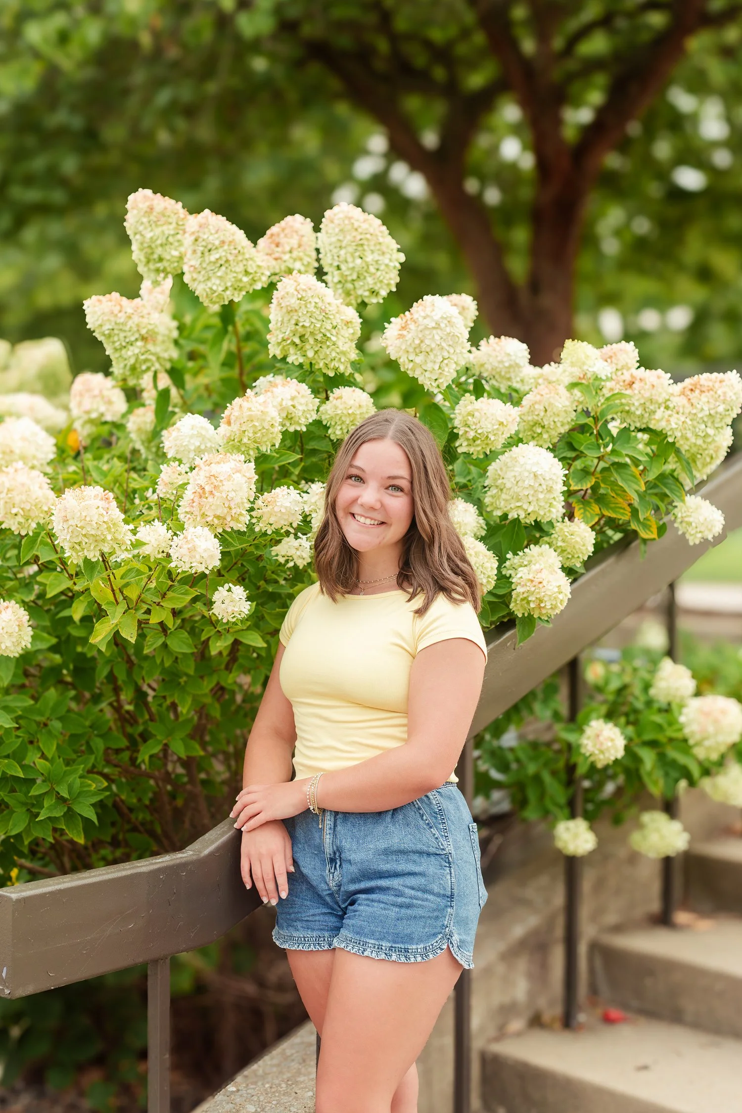 Young woman standing beside white flowering shrubs in denim shorts.