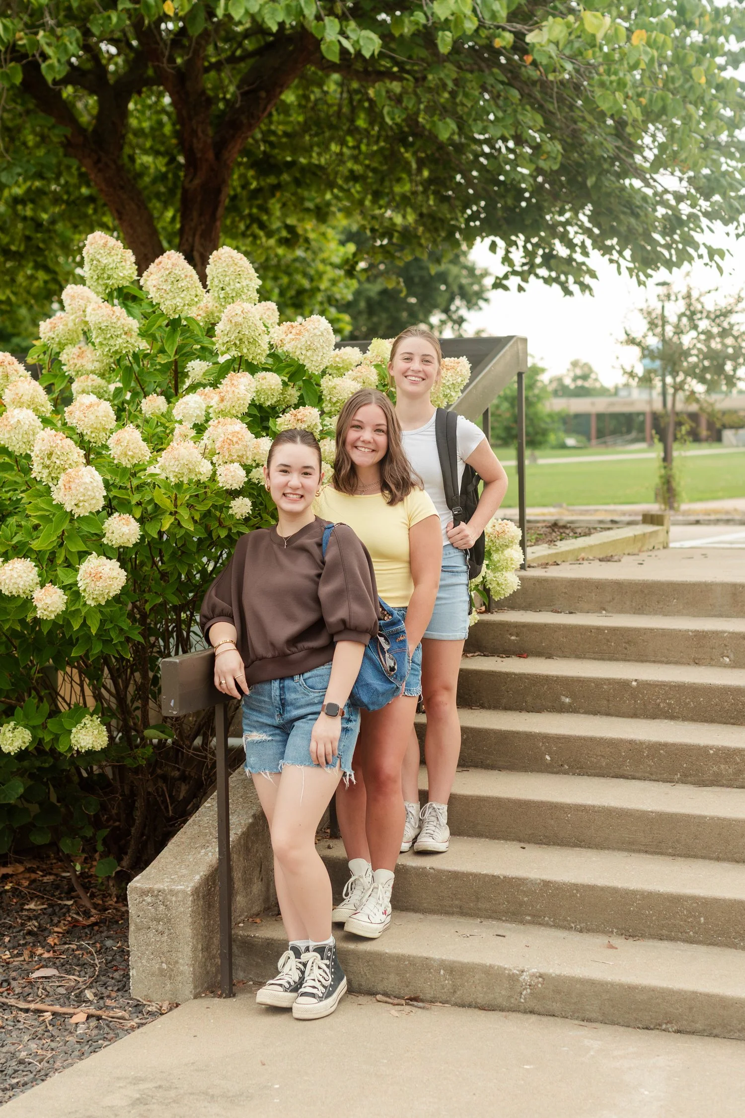 Three friends posing together on outdoor steps beside flowers.