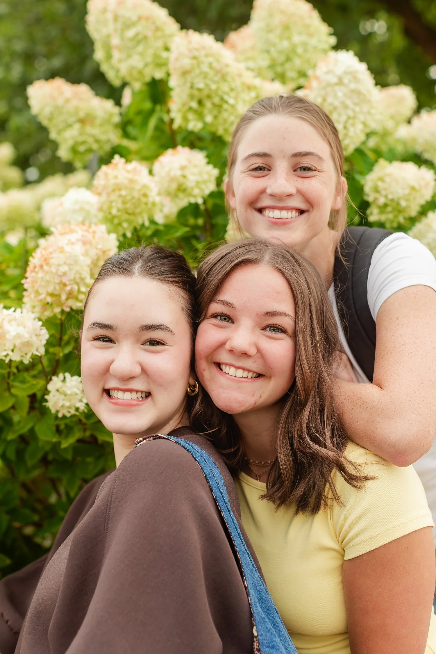 Three friends smiling close together with flowering shrubs behind them.
