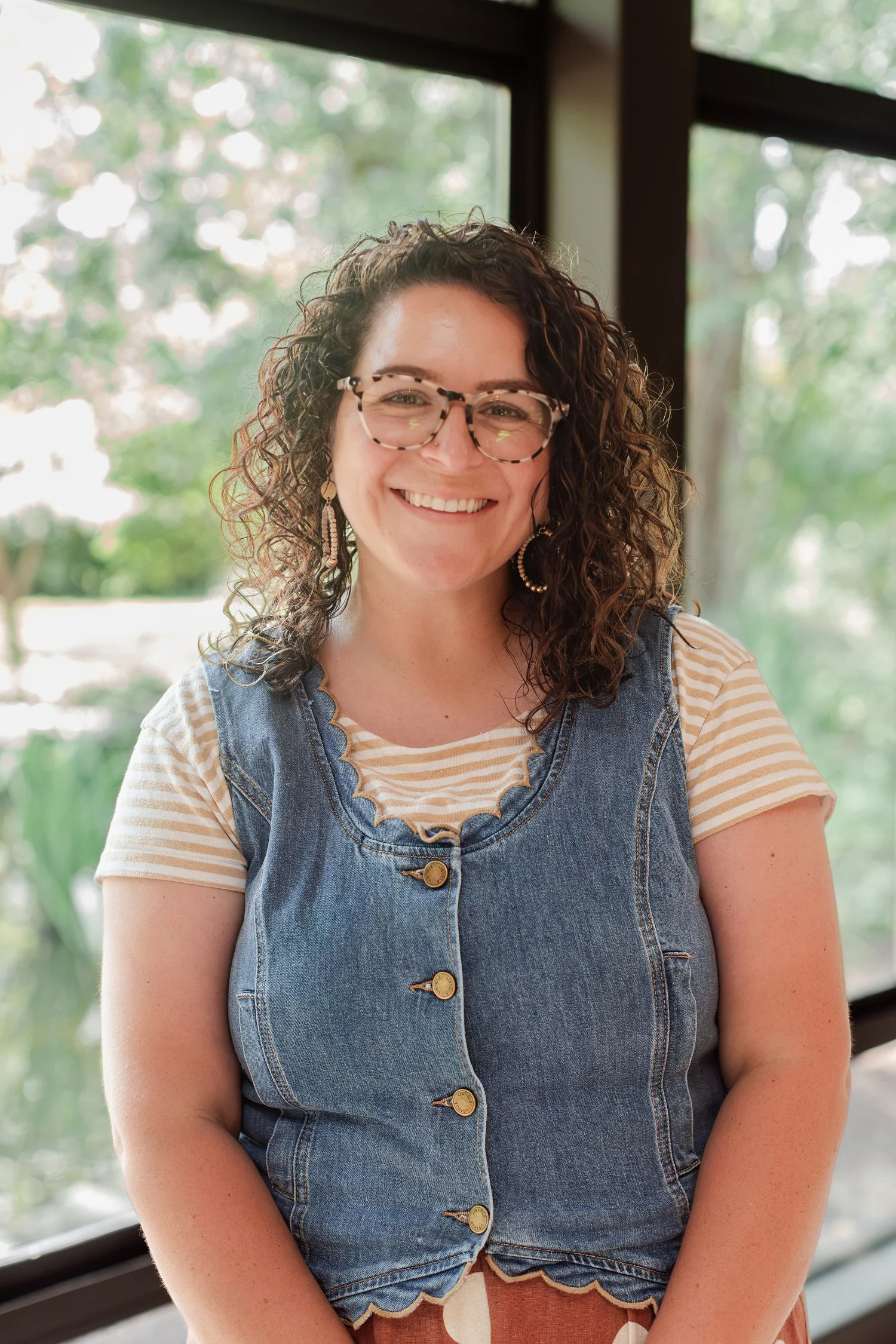 Real estate agent with curly hair and tortoiseshell glasses in a denim vest smiling by tall windows, real estate agent headshots in Bolivar, MO