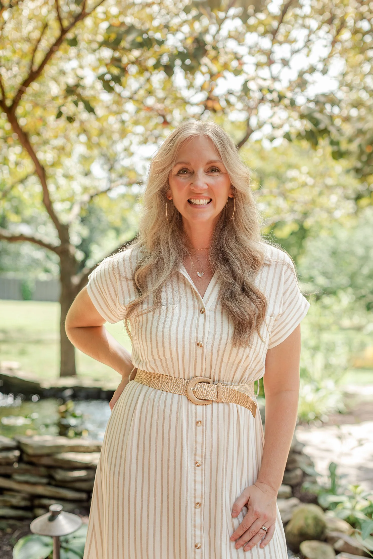Real estate agent in a cream striped shirt dress and woven belt standing in a garden by a pond, Stellar Real Estate Co. headshots in Bolivar, MO