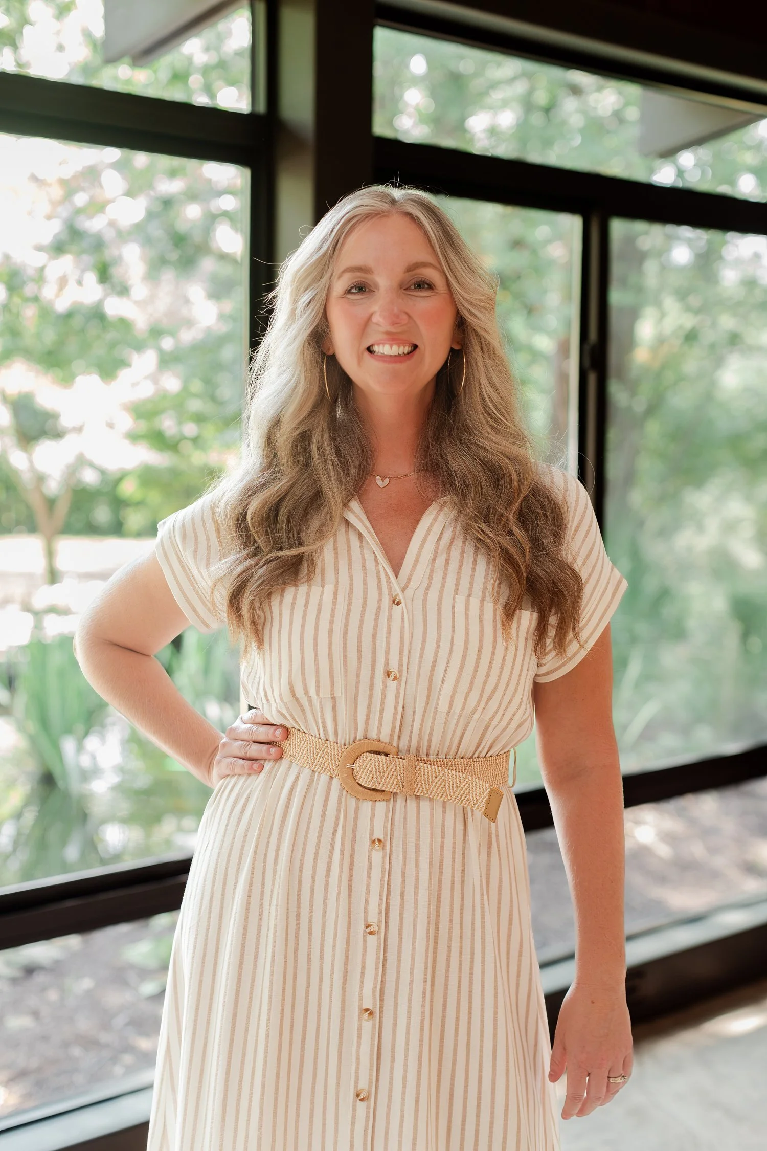 Real estate agent in a cream striped dress by floor to ceiling windows with trees outside, real estate headshots in Bolivar, MO