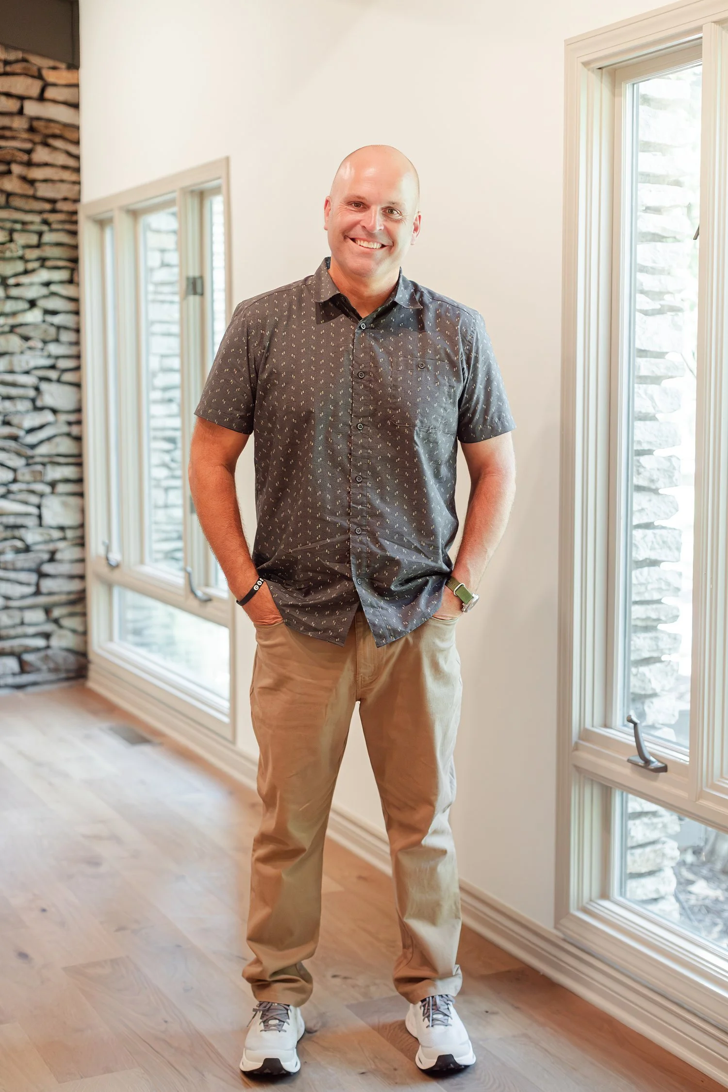 Real estate agent in a patterned short sleeve shirt and khakis standing between tall windows and a stone wall, full length headshot in Bolivar, MO