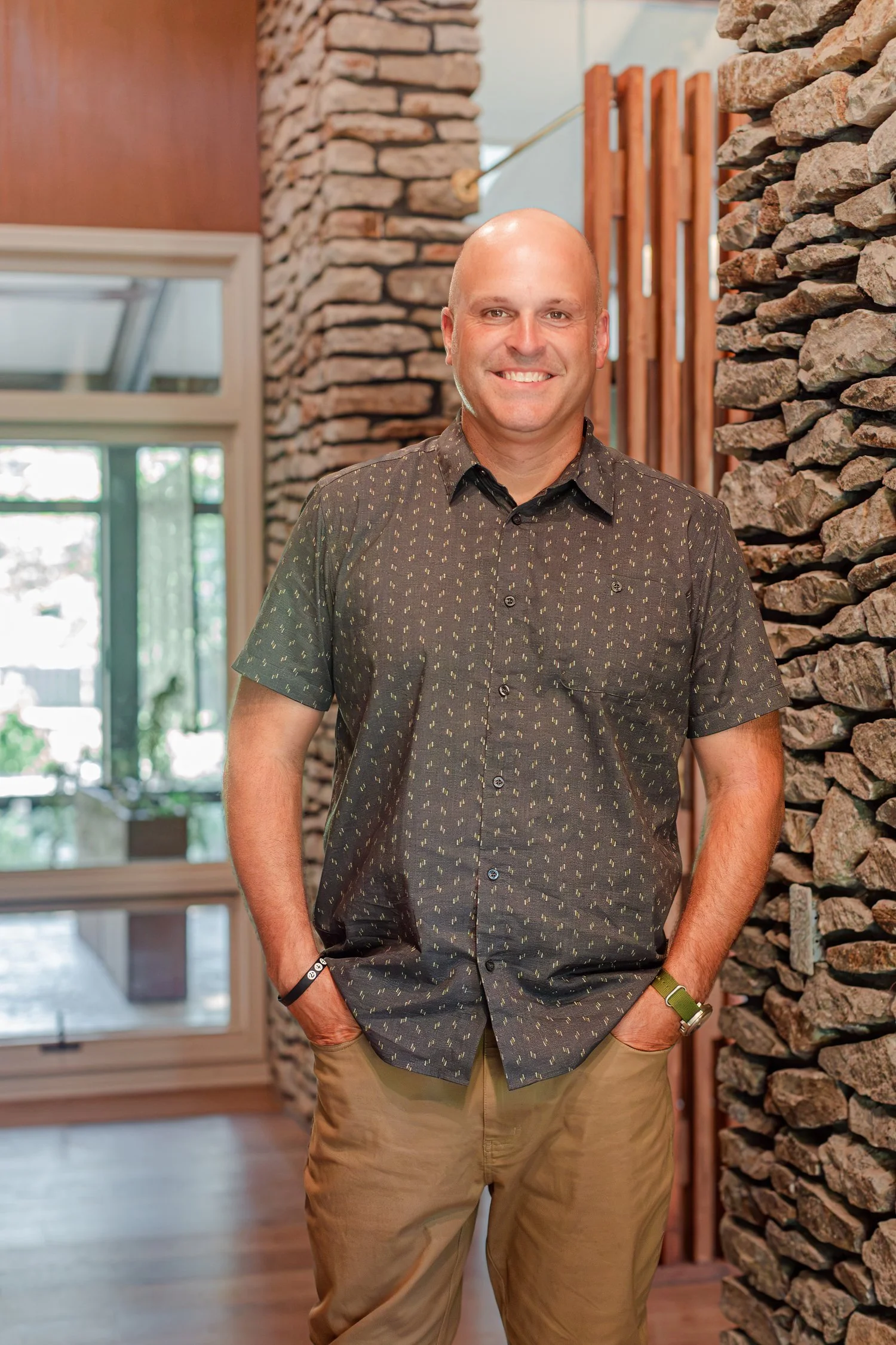 Real estate agent in a patterned button up shirt with hands in pockets beside a stone chimney, realtor headshots in Bolivar, MO
