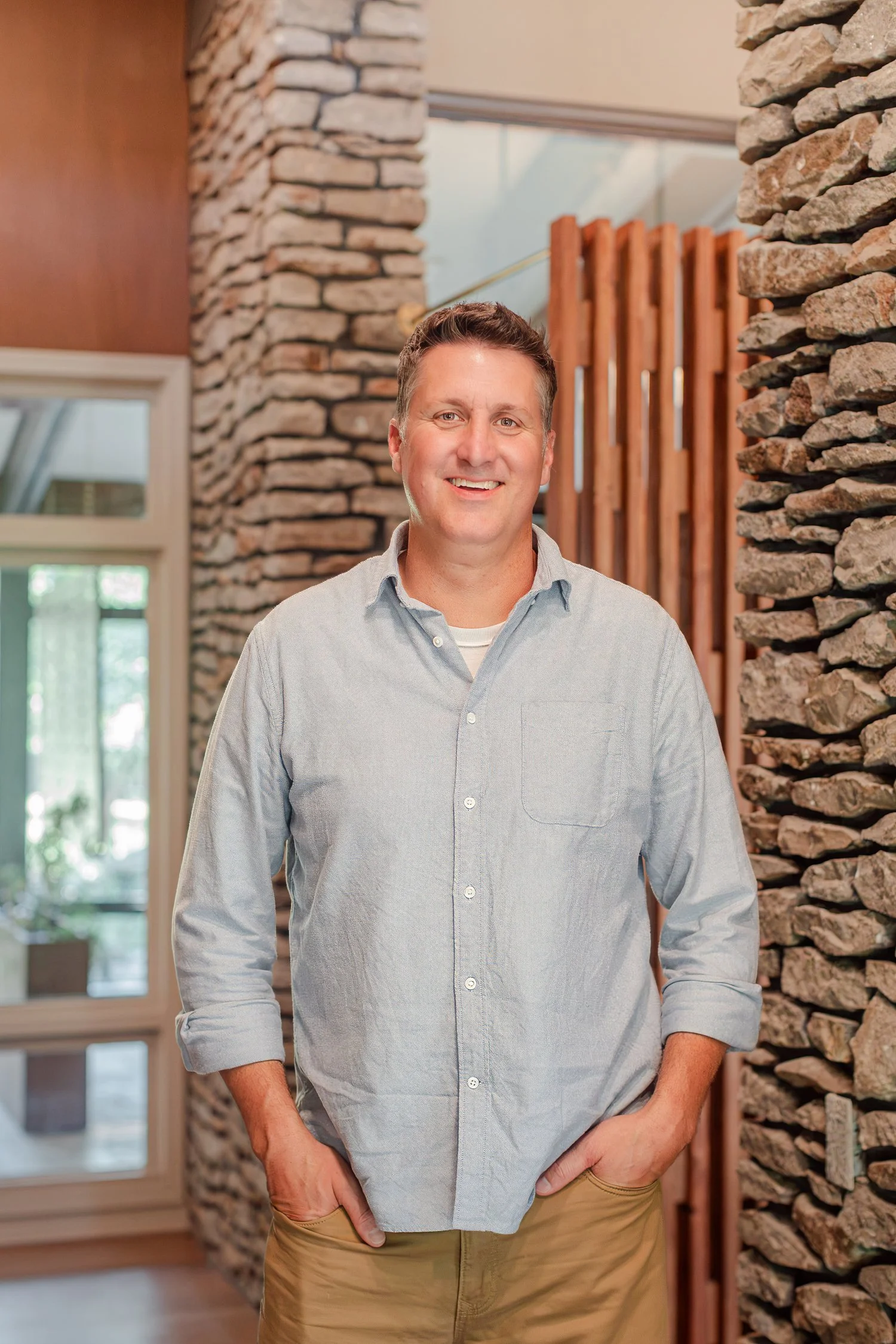 Real estate agent in a chambray shirt and khakis beside a stone chimney and wood slat screen, headshots in Bolivar, MO