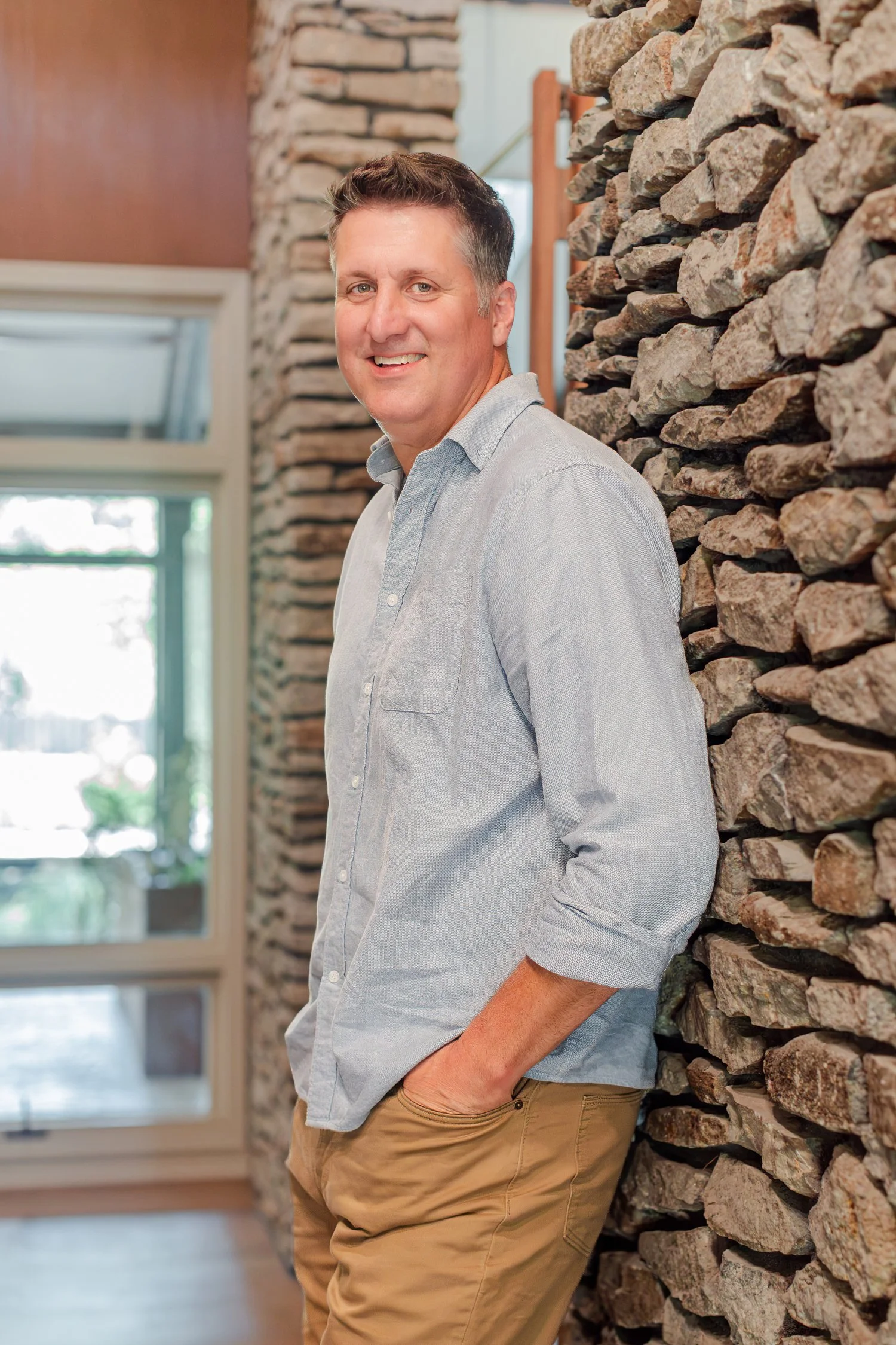 Real estate agent in a chambray shirt leaning against a stone wall, realtor headshots in the 417