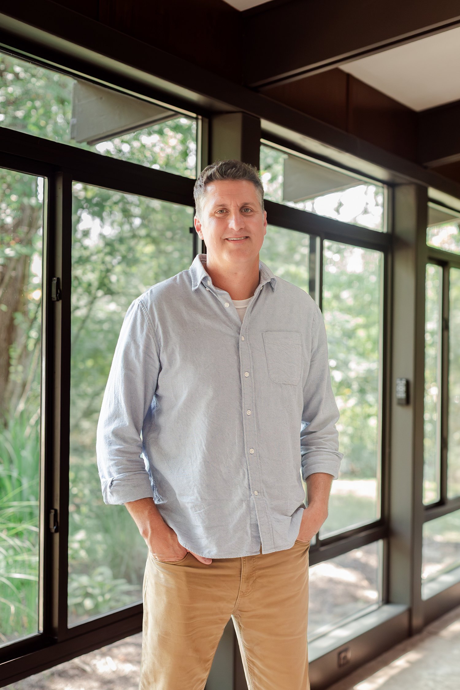 Real estate agent in a chambray shirt standing in a glass hallway with trees outside, team headshots in Bolivar, MO