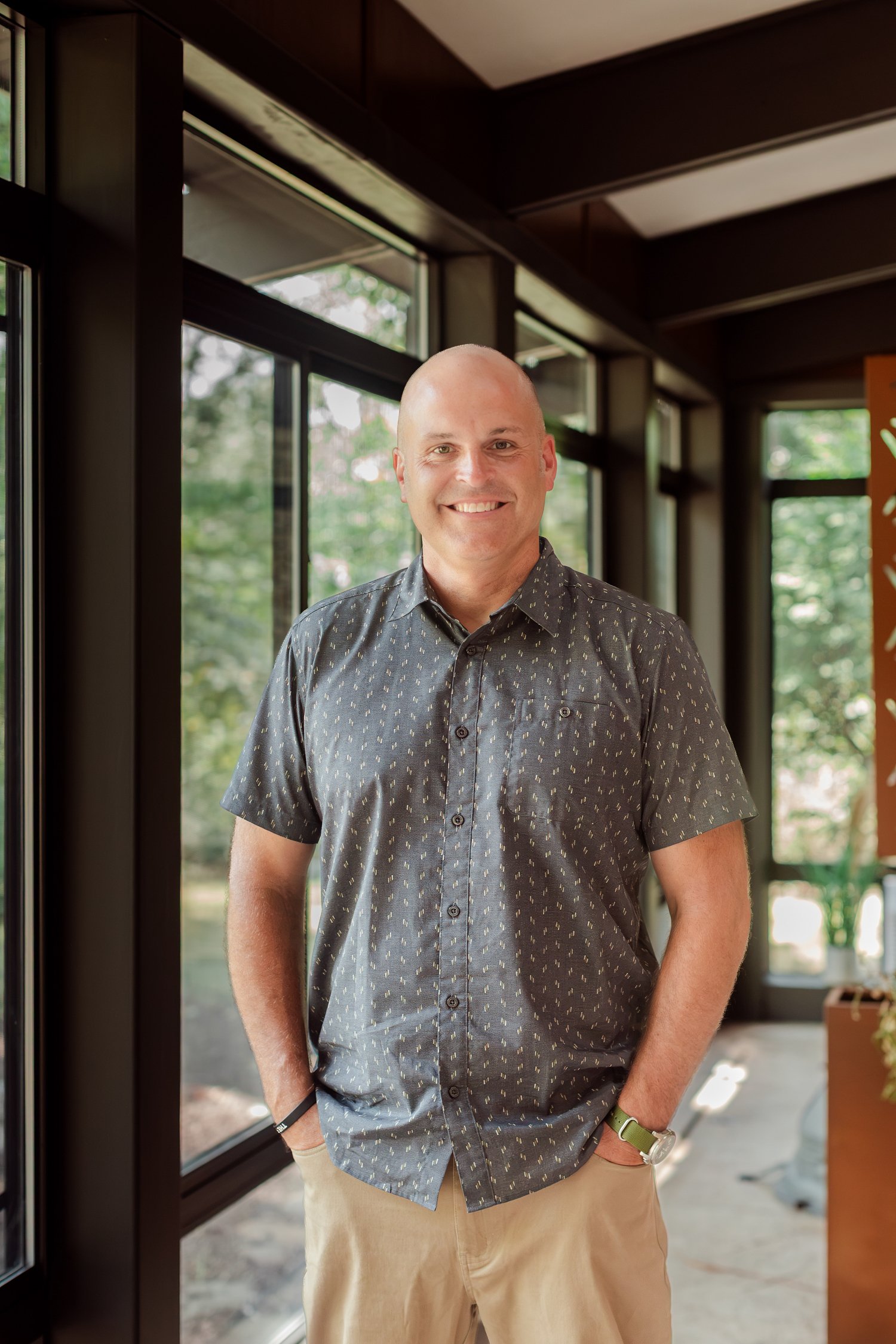 Real estate agent in a patterned shirt smiling in a glass hallway with dark window frames, Stellar Real Estate Co. headshots in Bolivar, MO