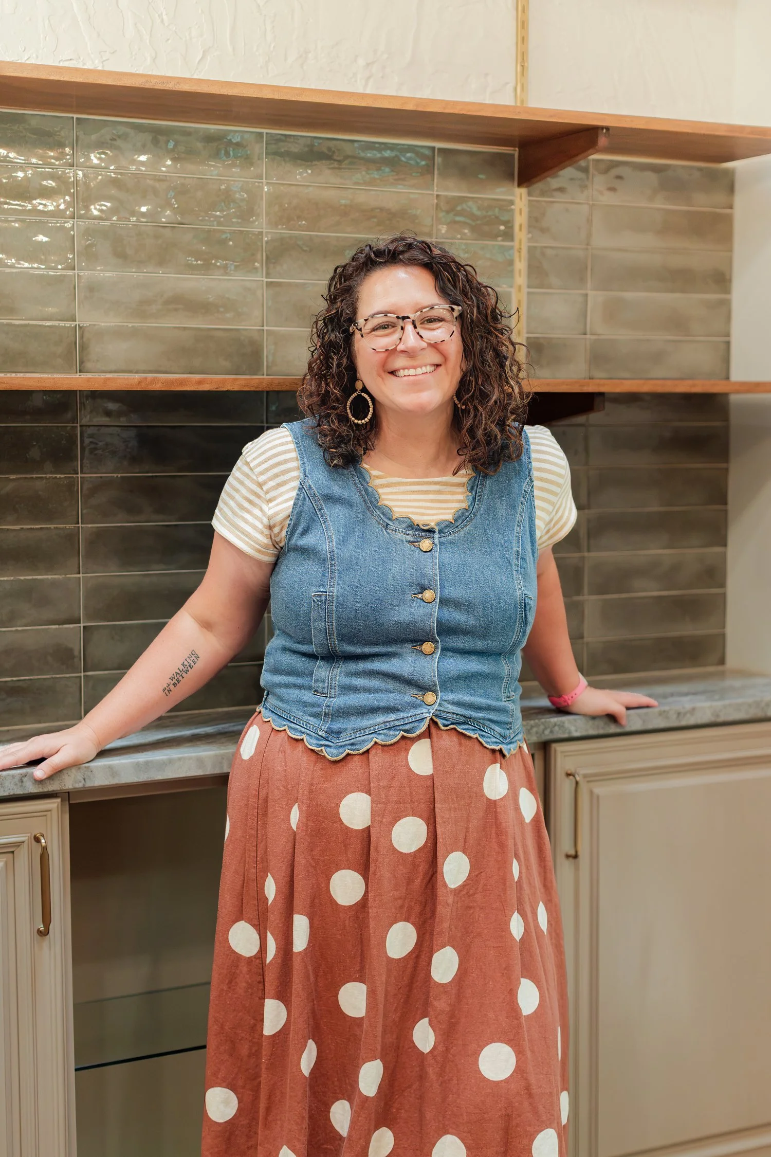 Real estate agent in a denim vest and rust polka dot skirt leaning on a kitchen counter with green tile behind, brand photos in Bolivar, MO