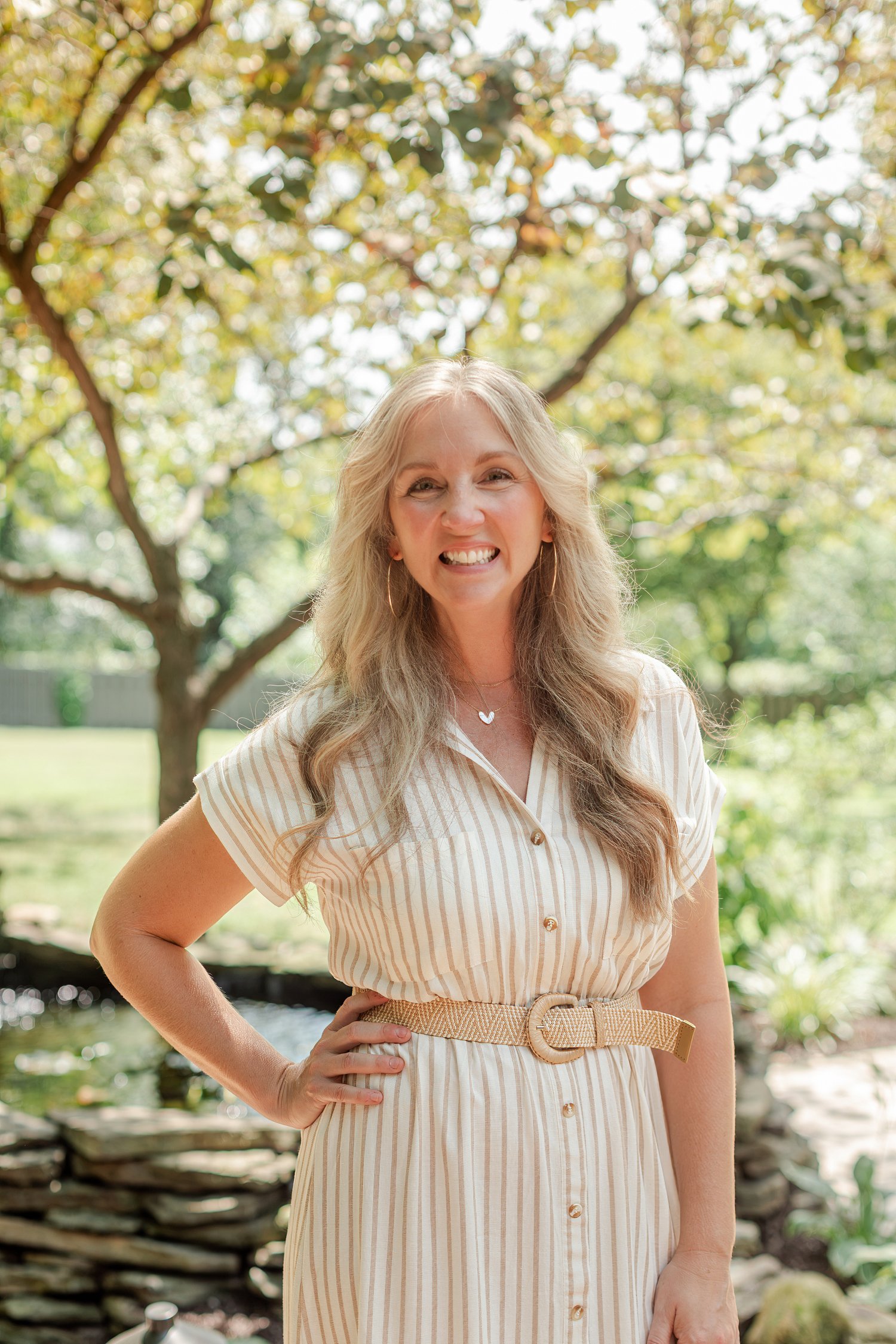 Real estate agent in a cream striped shirt dress with hands on hips under the trees by the pond, headshots in Bolivar, MO