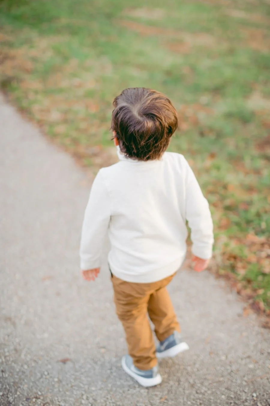 Young child in a cream sweater walking away along a path beside grass.